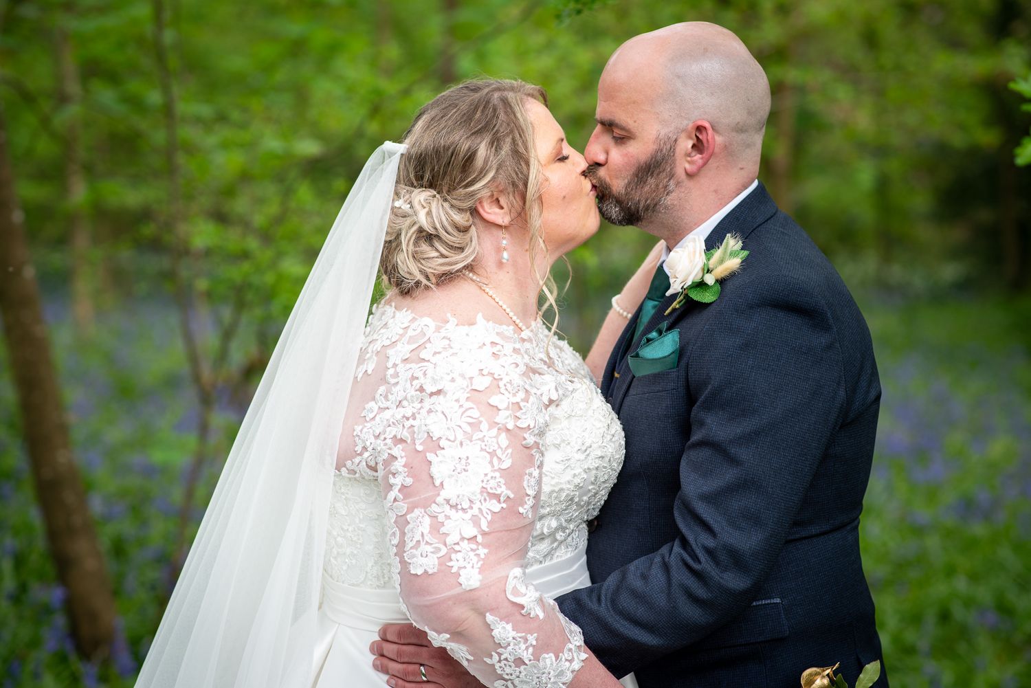 Bride and groom sharing romantic kiss in bluebell woodland, bride wearing lace wedding dress and veil.