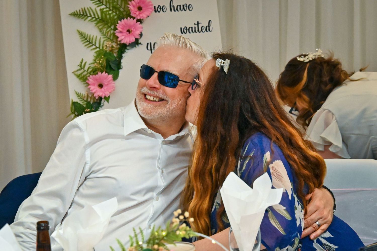 A woman kisses a smiling man wearing sunglasses at a wedding reception near a floral sign.