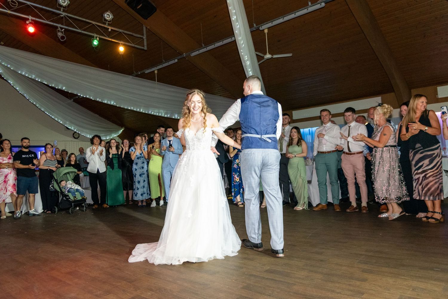 Bride in white gown and groom share first dance on venue floor surrounded by applauding wedding guests.