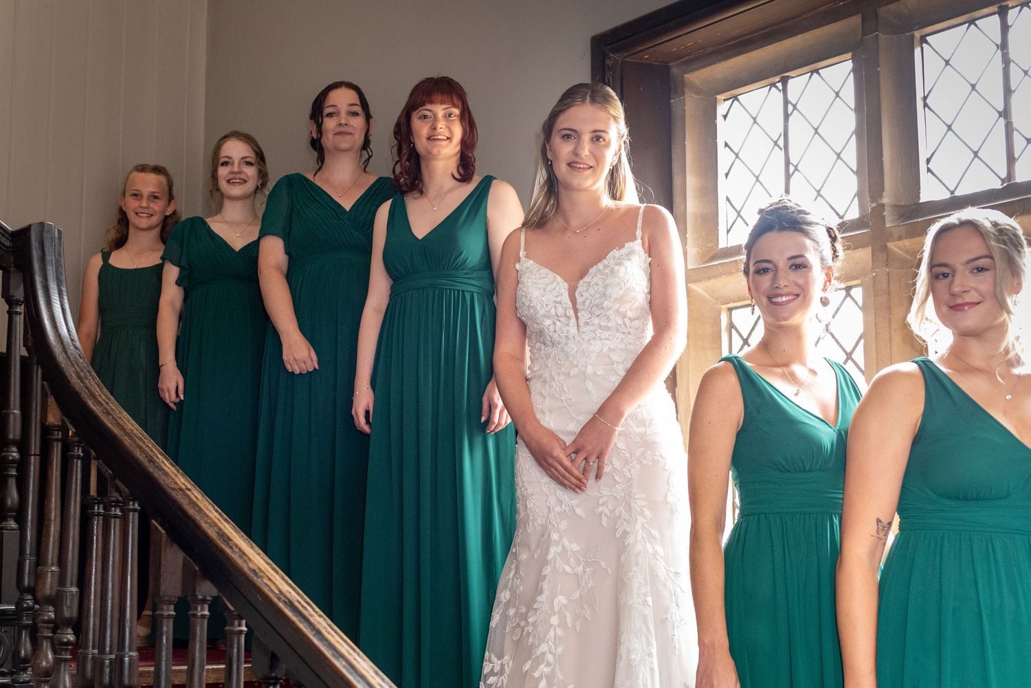 Bride in white lace gown poses with six bridesmaids in emerald green dresses on an elegant staircase.