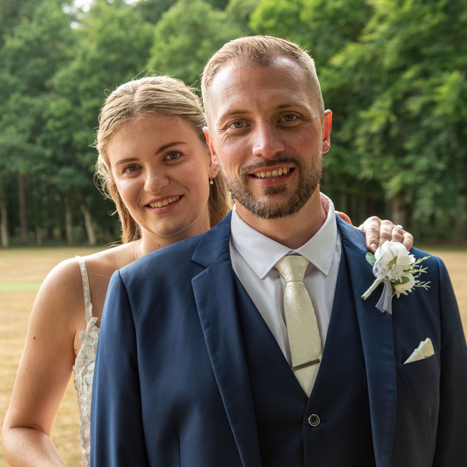 A smiling couple in wedding attire poses outdoors, man in navy suit with boutonniere, woman in white dress.