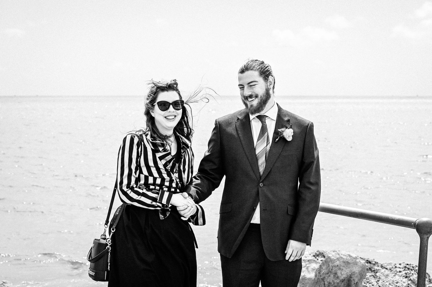 Black and white photo of a smiling couple holding hands outdoors near the ocean, man in suit with boutonniere.