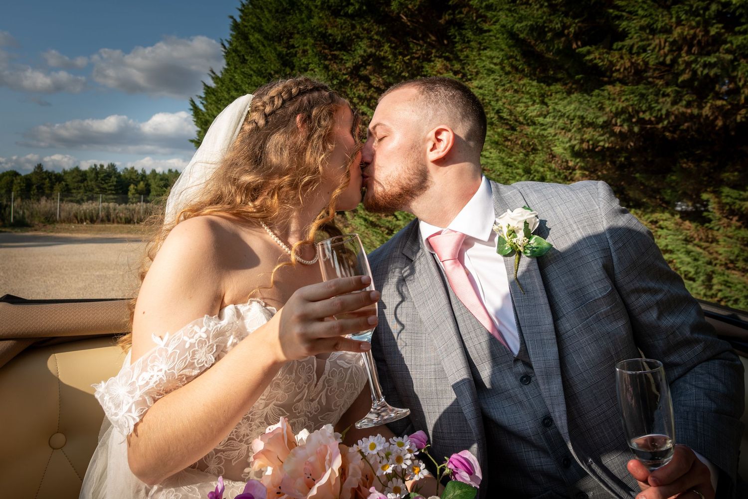 Bride and groom kissing while holding champagne glasses in vintage car on wedding day.