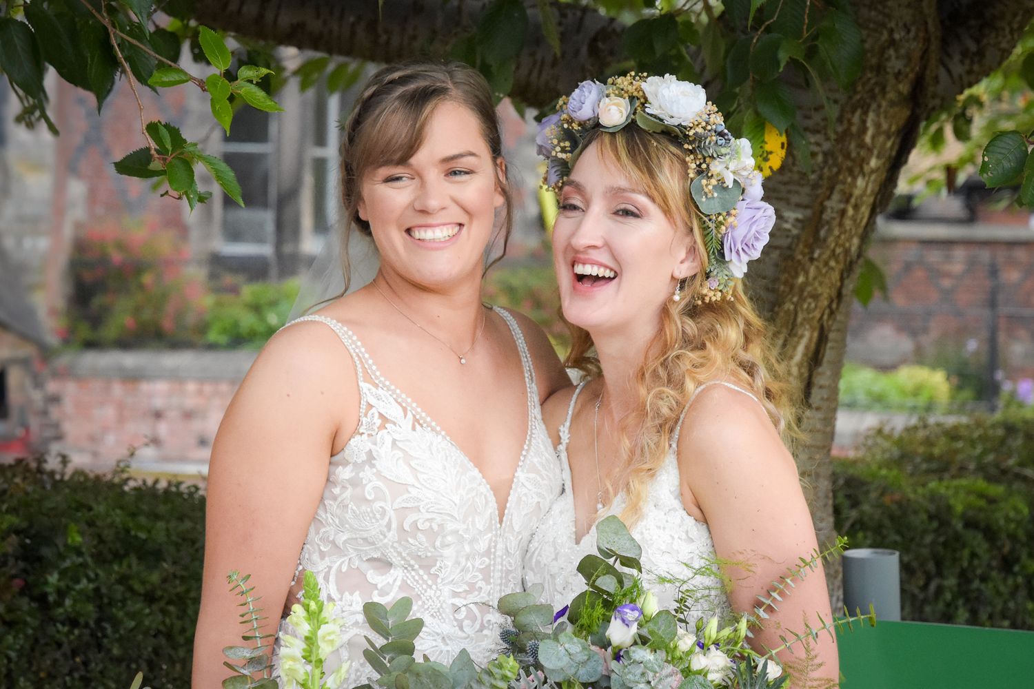 Two brides in white wedding dresses smile joyfully, one wearing a floral crown, holding a bouquet outdoors.