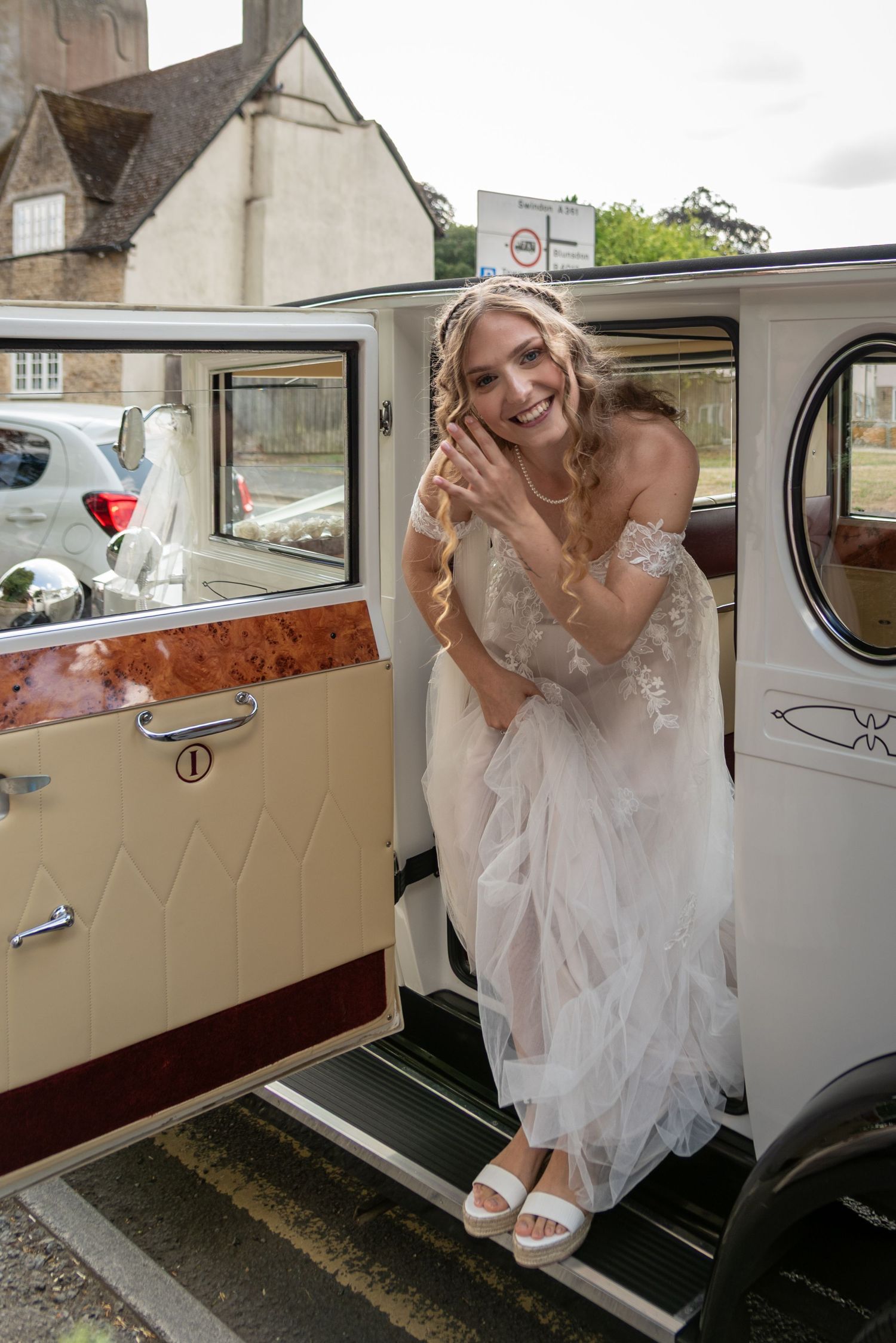 Smiling bride in white lace gown stepping out of a vintage cream wedding car, wearing white heels and a hair accessory.