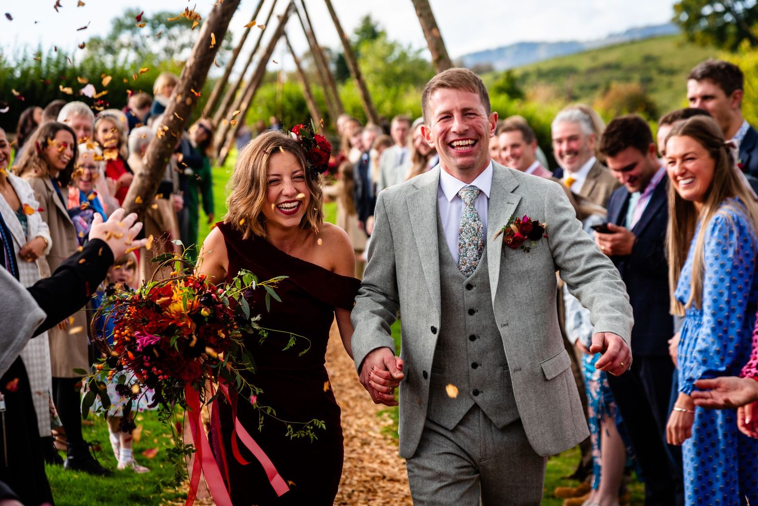 Joyful bride in burgundy dress and groom in grey suit walking through confetti shower at outdoor wedding ceremony.