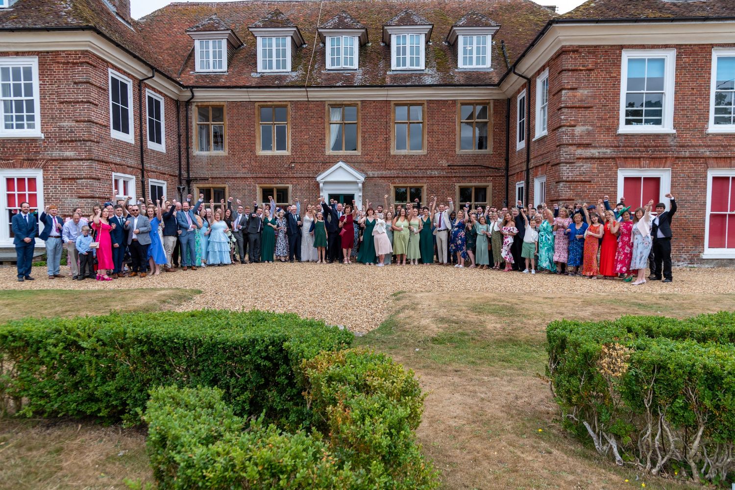 Large wedding group photo outside a grand red brick English manor house with manicured hedges.