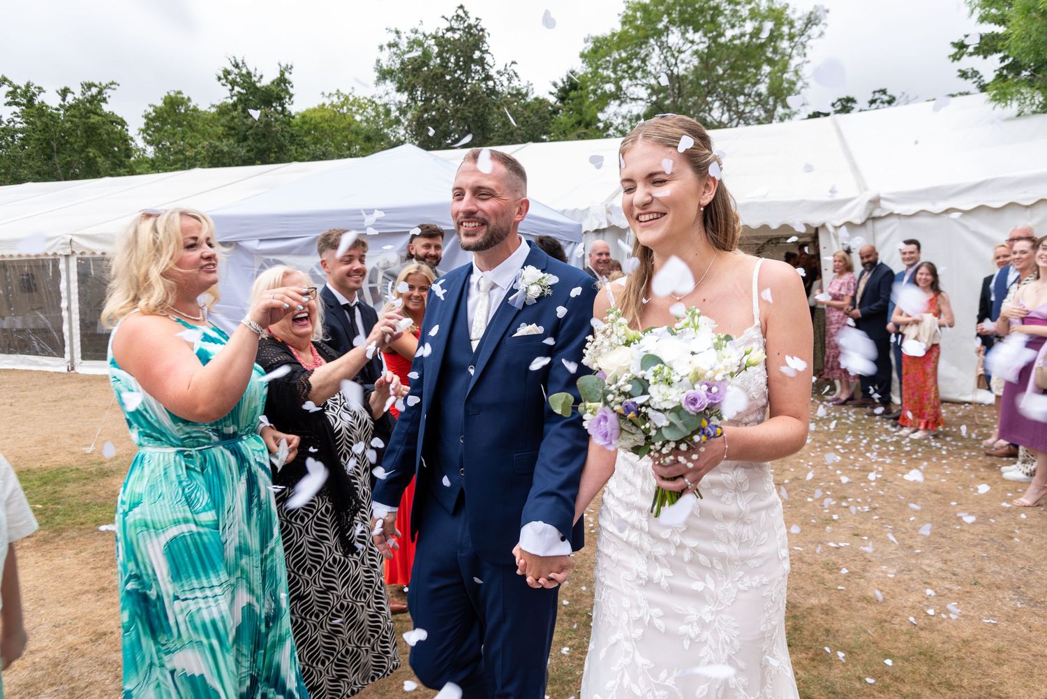 Bride and groom walk through confetti thrown by cheering guests outside a white marquee wedding tent.