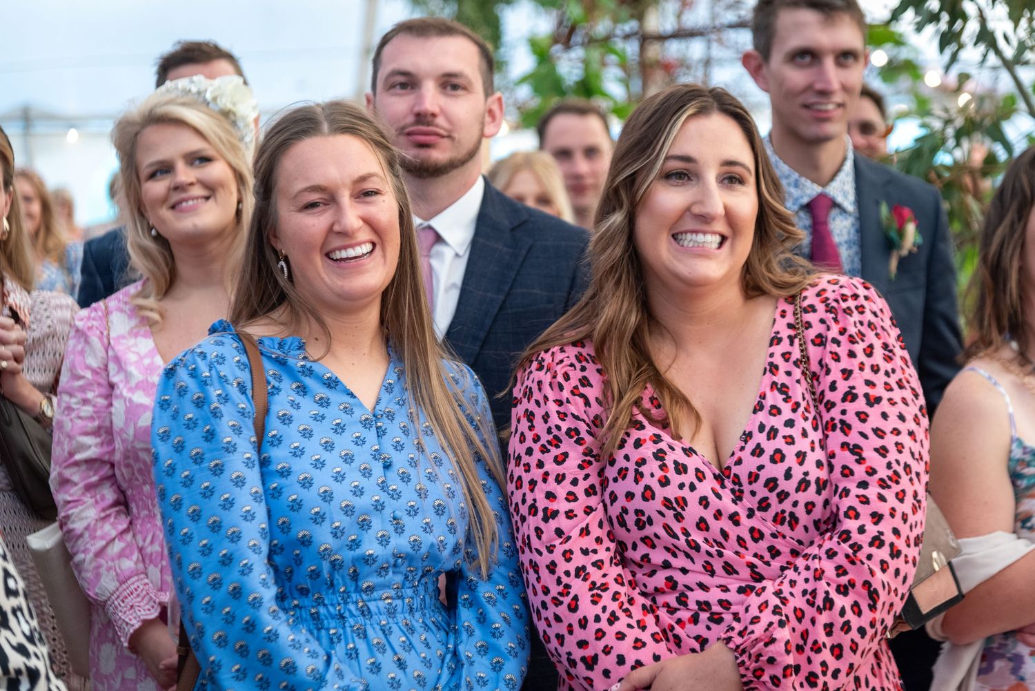 Two smiling women in floral dresses at a wedding celebration with guests in background.