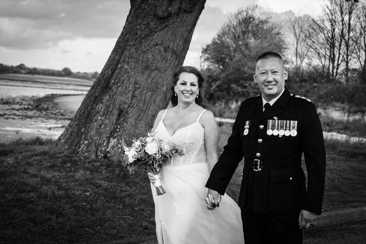 Black and white wedding photo of bride in white gown and groom in military uniform holding hands outdoors.
