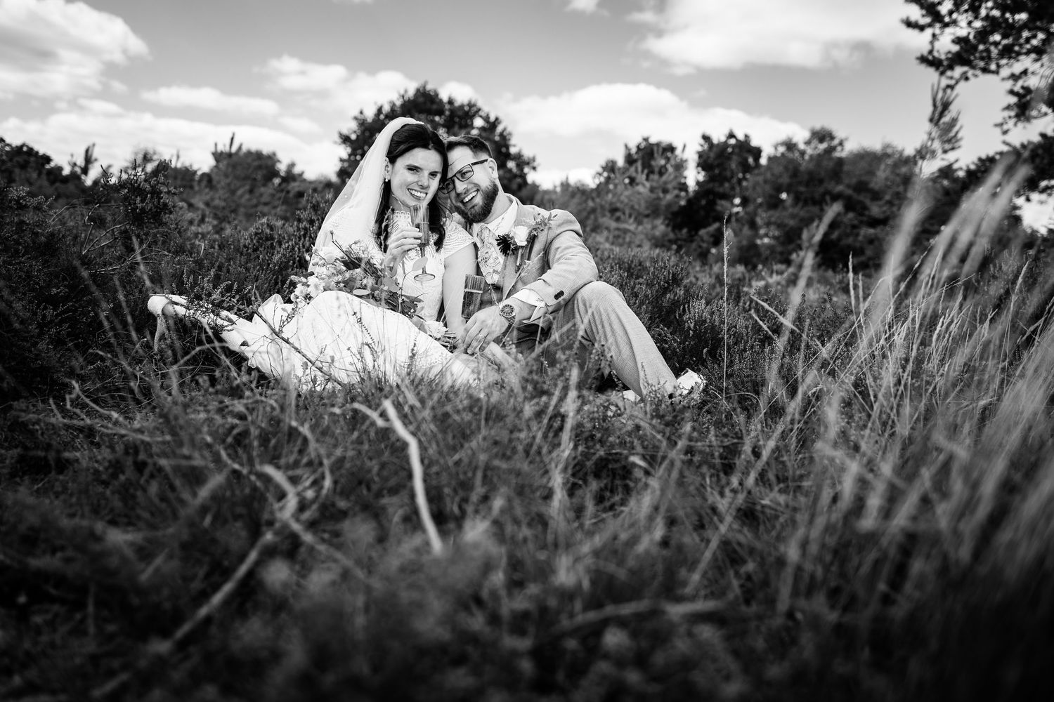 Black and white photo of a bride and groom sitting together in tall grass, smiling on their wedding day.