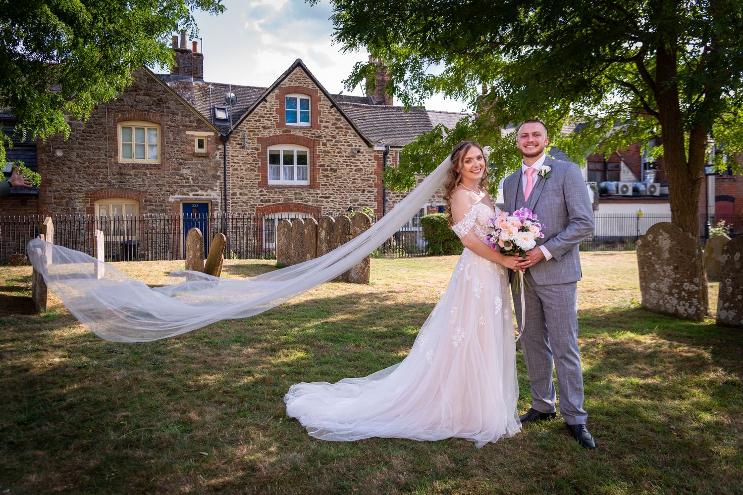 Bride and groom posing outdoors by a stone church, bride wearing a flowing veil and holding a bouquet.
