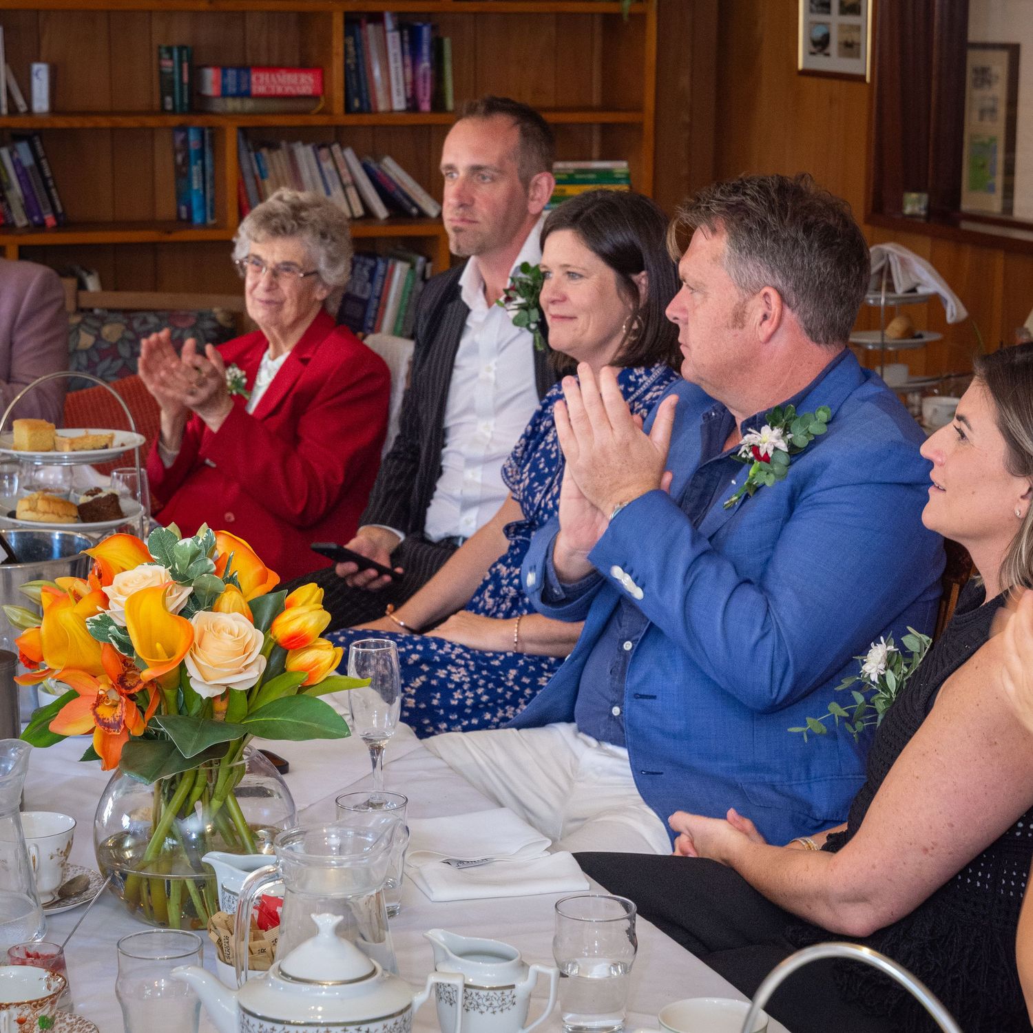 Guests seated at a decorated wedding reception table with yellow tulip centerpieces in a room lined with bookshelves.