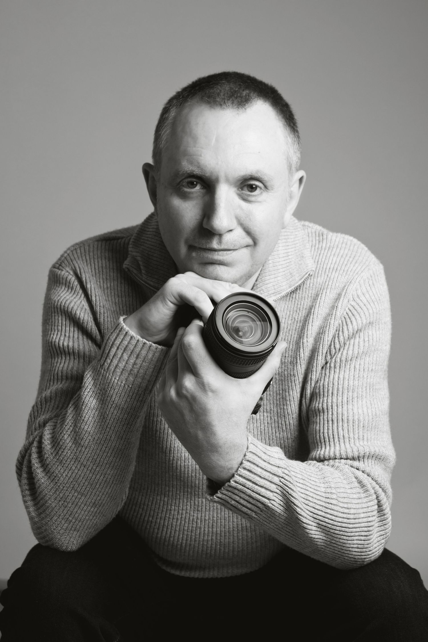 Black and white portrait of a middle-aged man holding a camera lens, wearing a knit sweater, smiling at the camera.