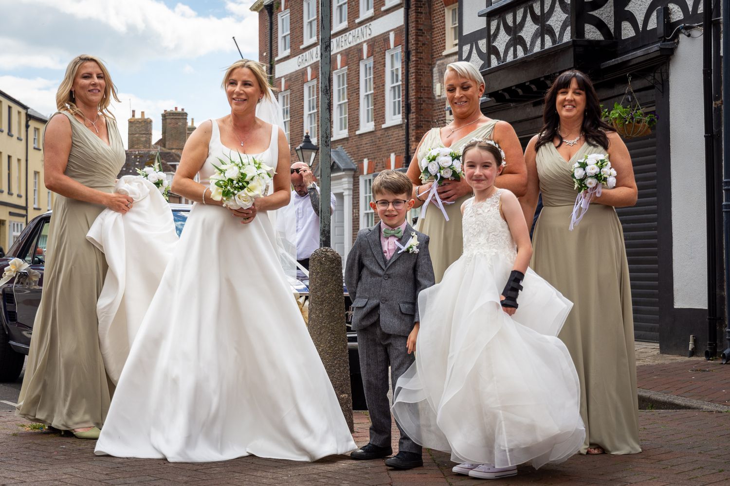 Bride in white gown with bridesmaids in sage green dresses, flower girl, and page boy on a sunny street.