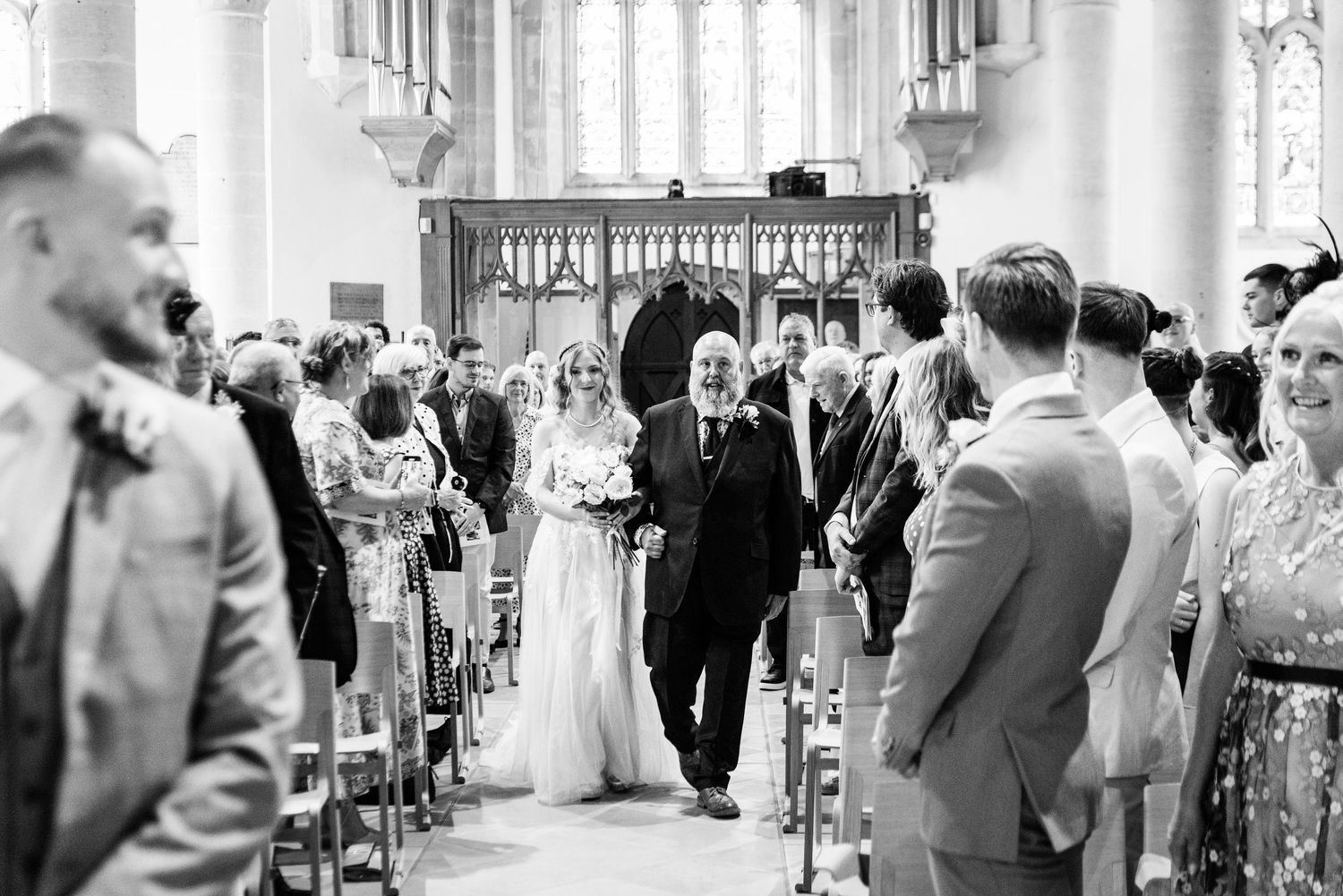 Bride walks down church aisle with escort in black and white wedding ceremony photo.