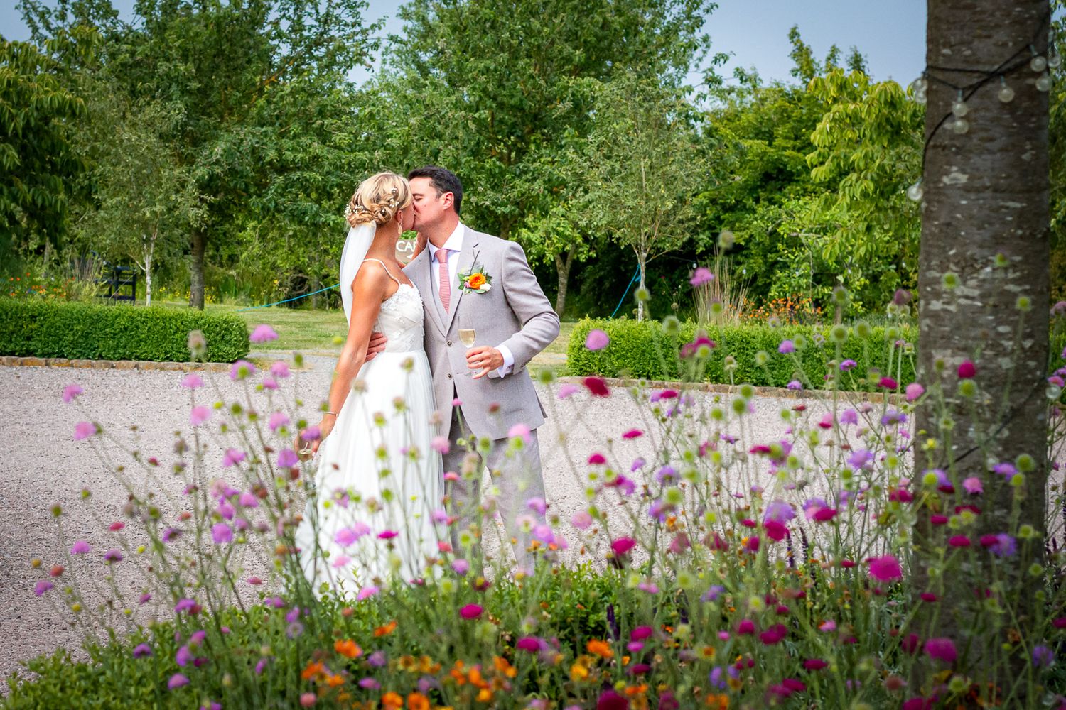 A bride and groom share a romantic kiss surrounded by colorful wildflowers in a lush garden on their wedding day.
