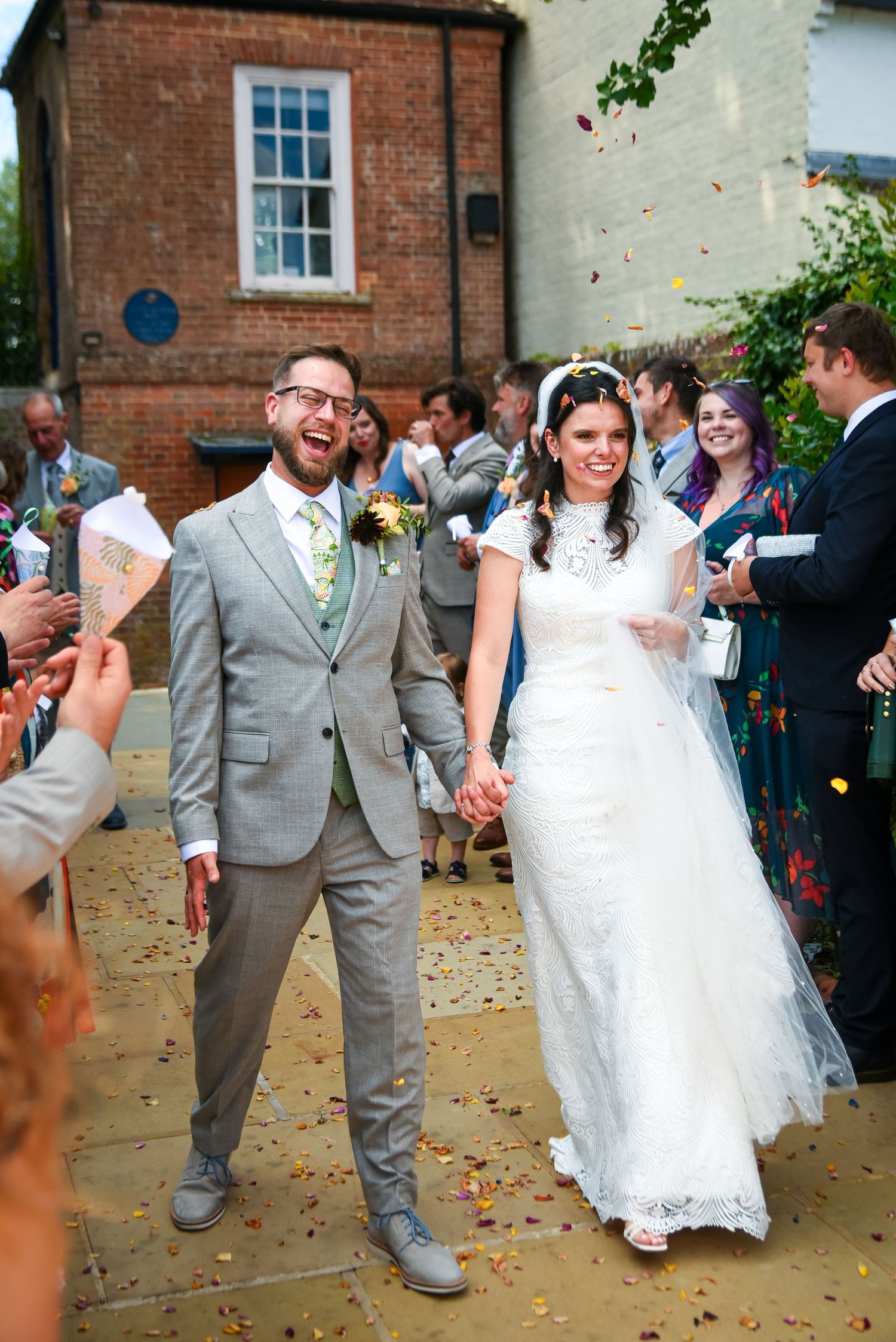 Joyful newlyweds walking through confetti shower outside venue, groom in grey suit, bride in lace wedding dress with veil.