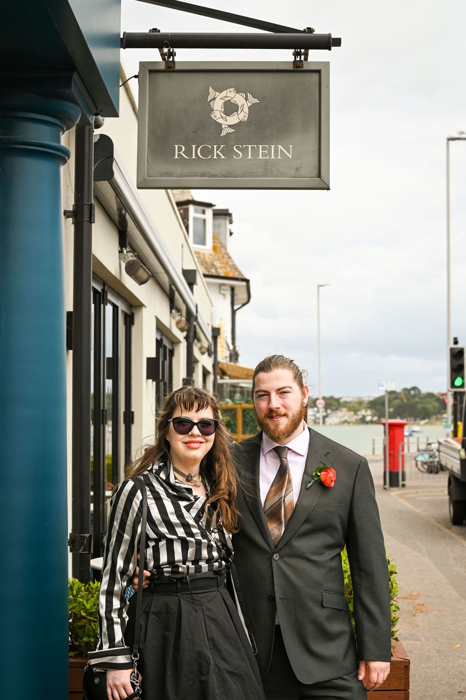 Couple posing outside Rick Stein restaurant, man in suit with boutonniere, woman in striped blouse, waterfront setting.