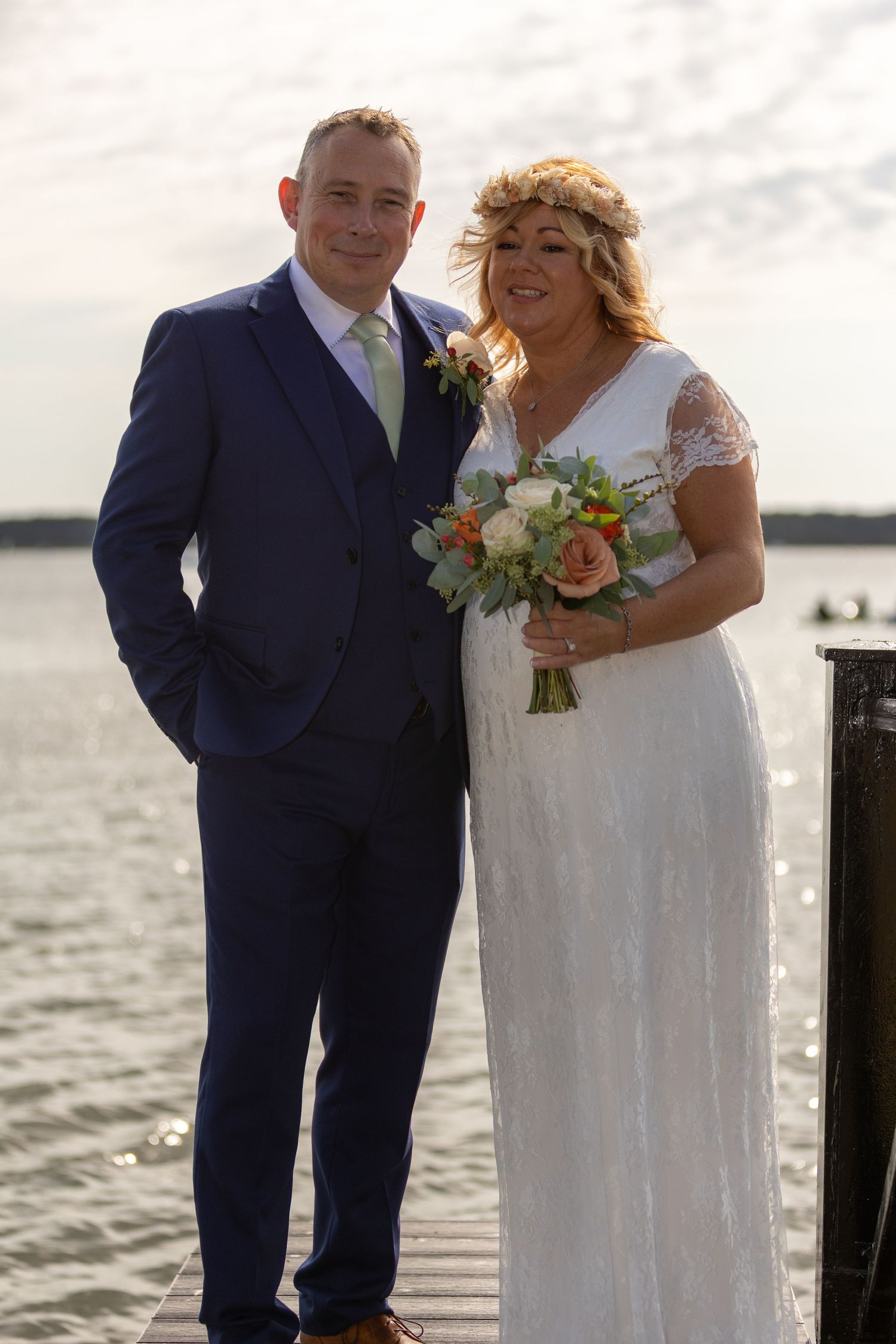 Bride and groom posing on a waterfront dock, bride in lace gown with floral crown holding bouquet, groom in navy suit.