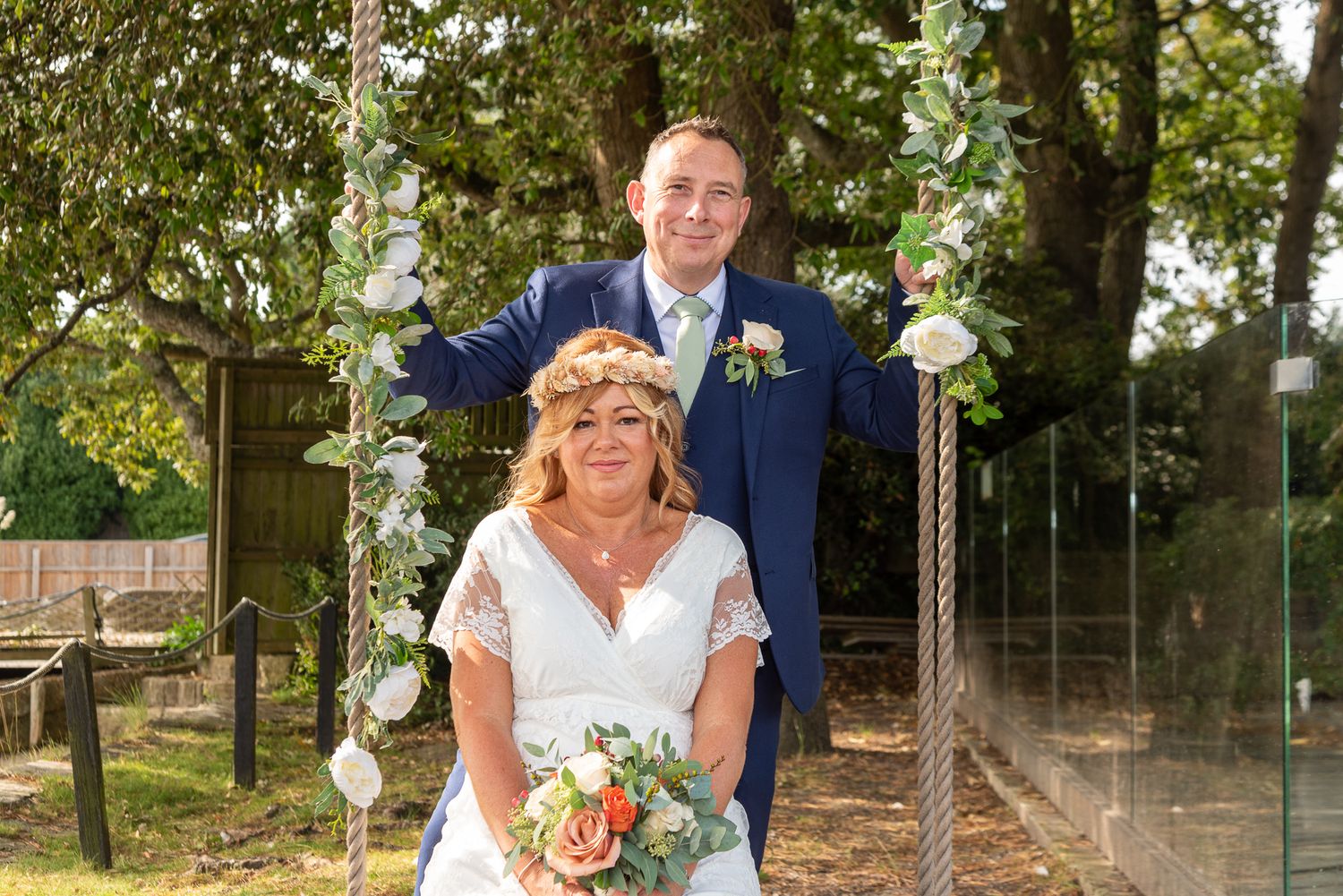 Bride and groom pose by a rope swing outdoors, bride holding a floral bouquet and wearing a flower crown.