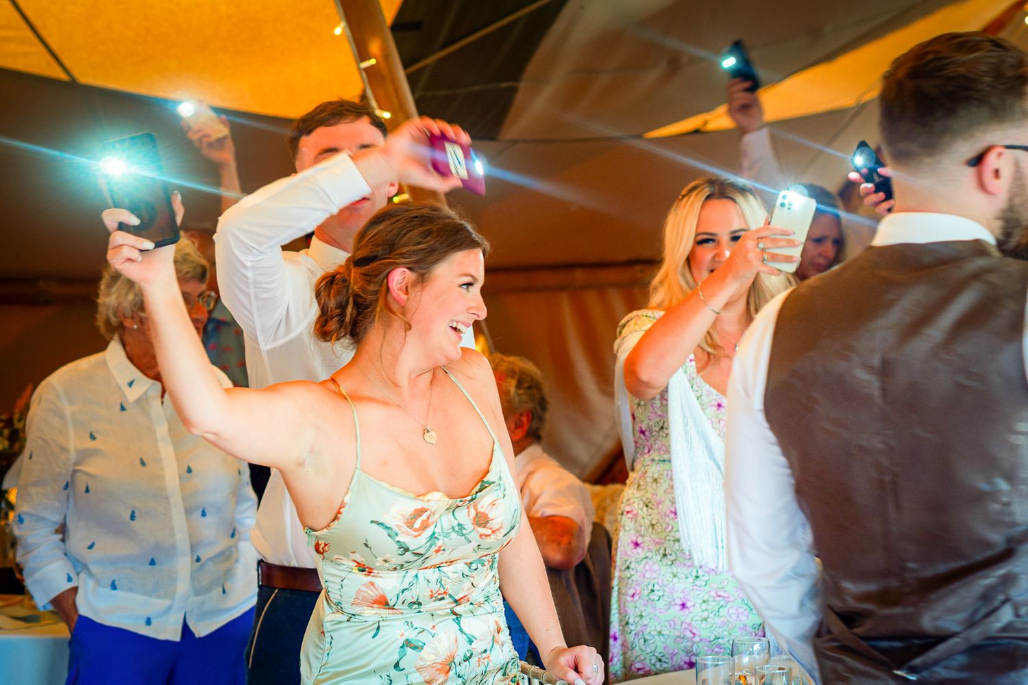 Guests dancing joyfully at a wedding reception, holding phones with flashlights on in a warmly lit tent venue.