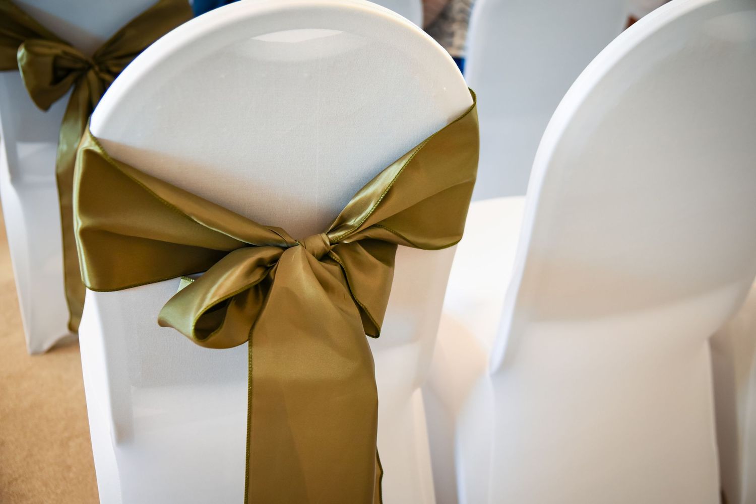 White wedding chair covers adorned with elegant gold satin ribbon bows at a formal event venue.