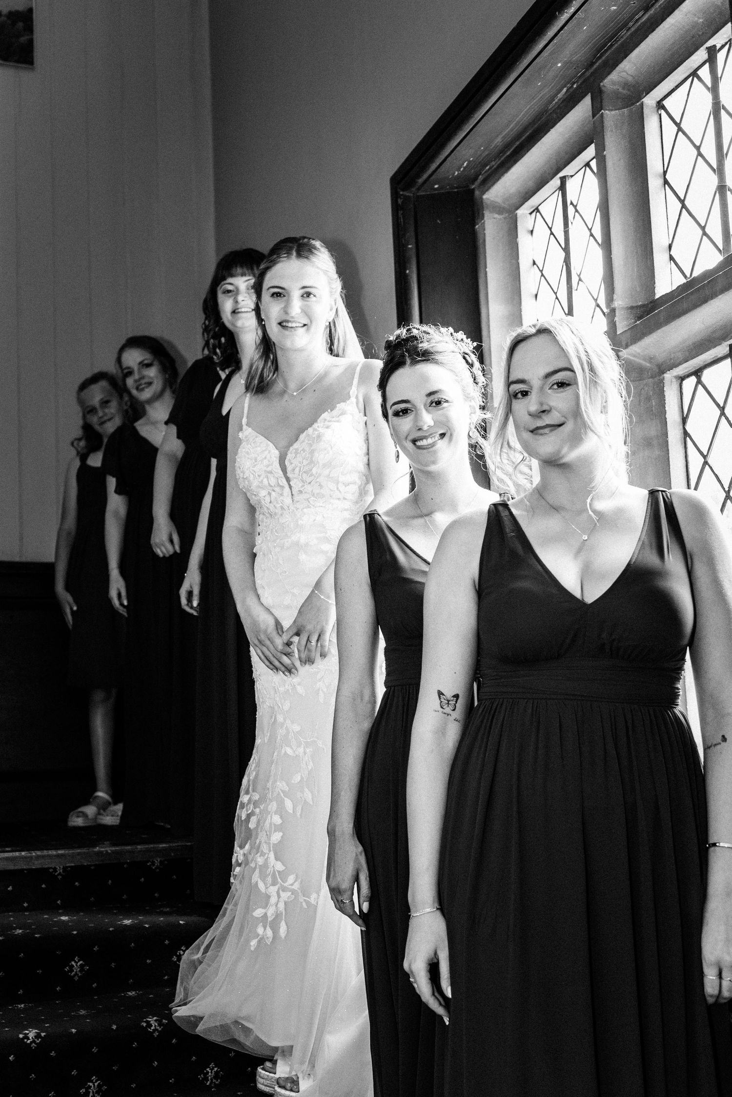 Bride in white lace gown poses with bridesmaids in black dresses near a window in black and white photo.
