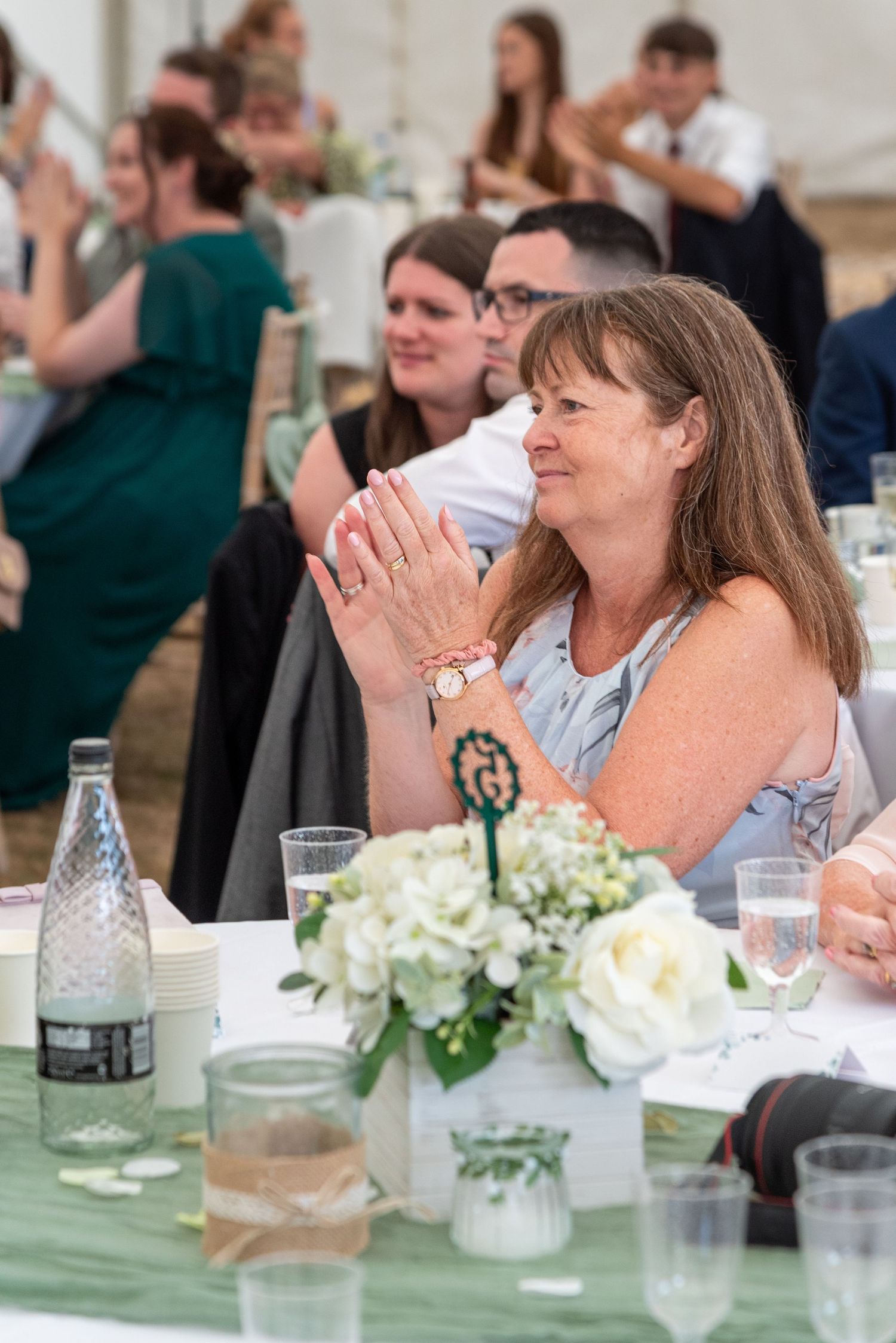 Wedding guest claps hands smiling at reception table with white floral centerpiece in decorated marquee.