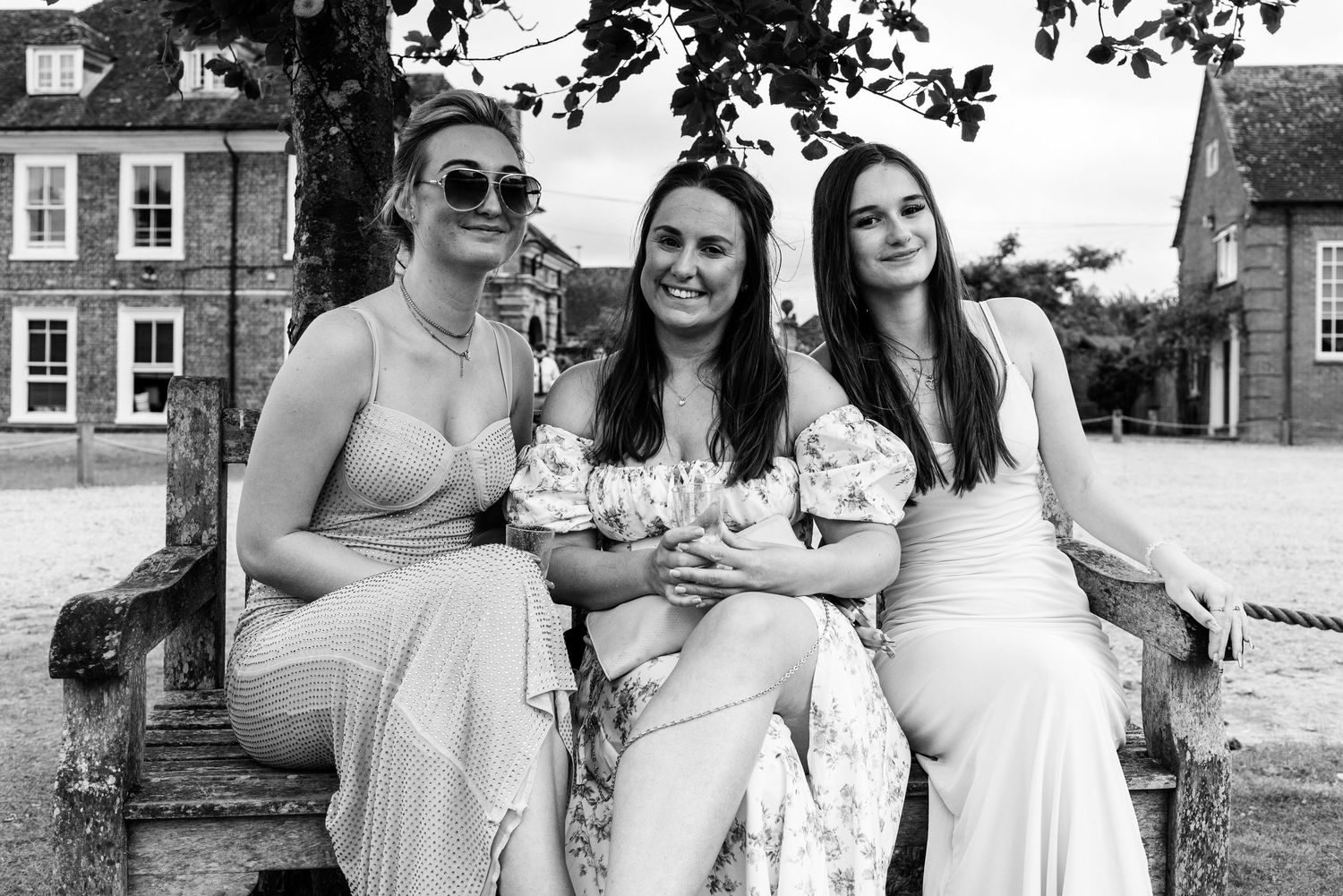 Three women in elegant dresses smile together on a wooden bench outdoors at a wedding reception.