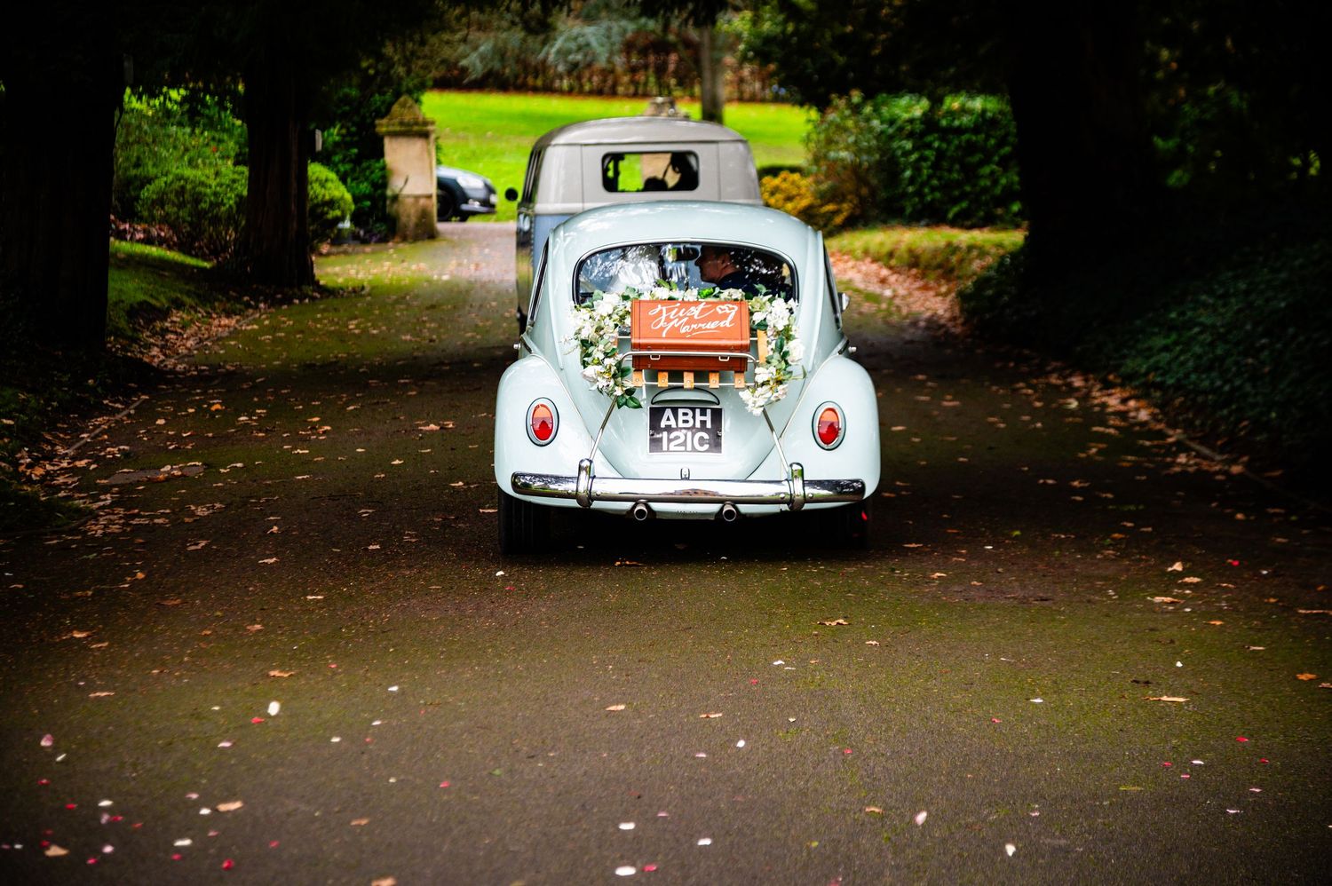 Vintage white VW Beetle wedding car decorated with flowers drives down a tree-lined path.