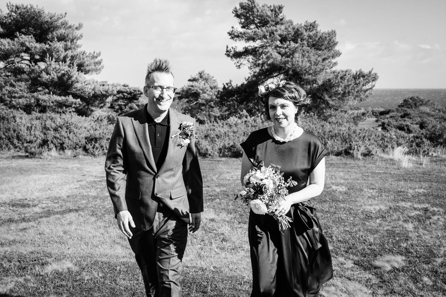 Black and white photo of a couple outdoors, man in suit and woman holding bouquet, trees in background.