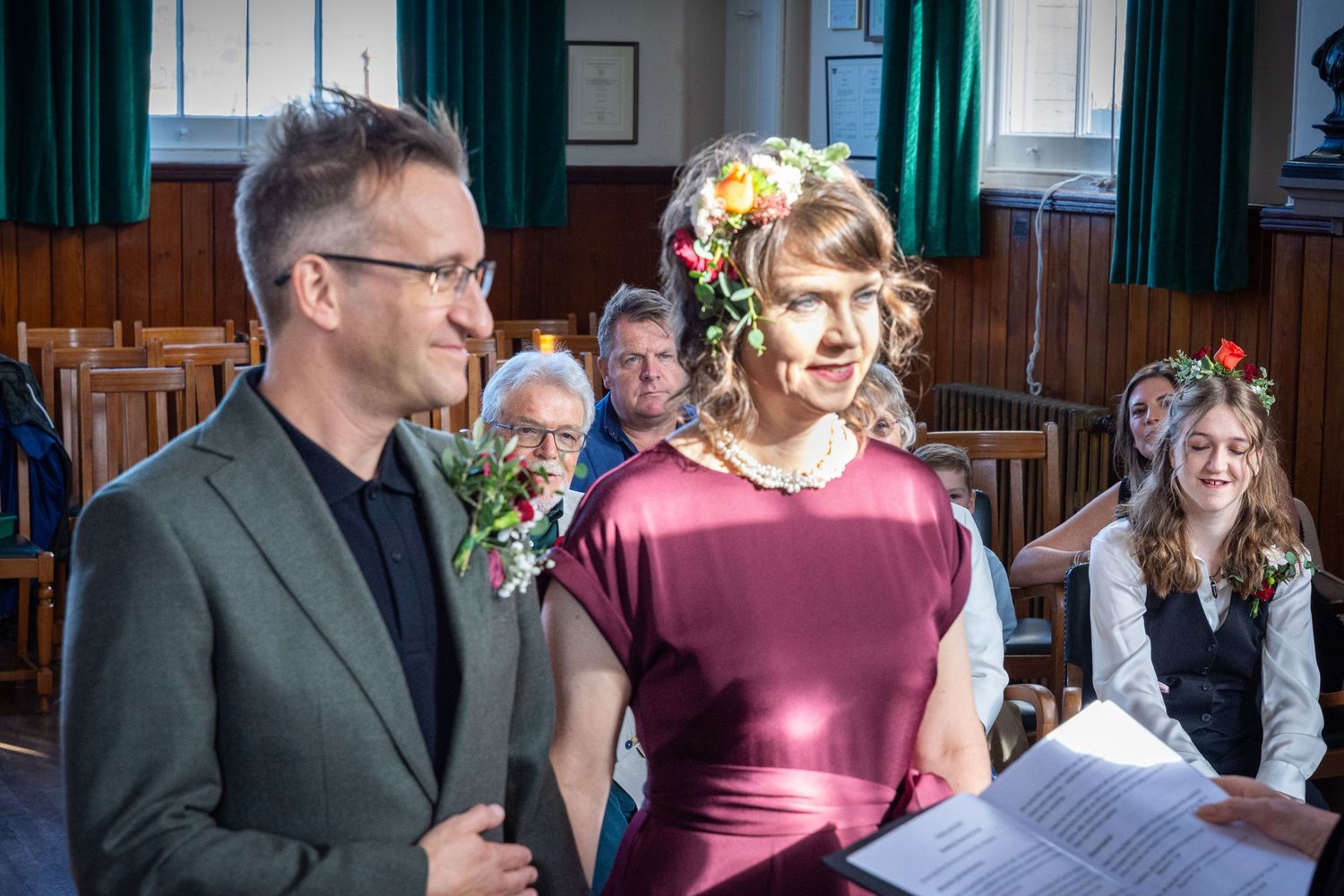 Couple smiling at wedding ceremony, bride wearing floral crown and burgundy dress, groom in gray suit holding bouquet.