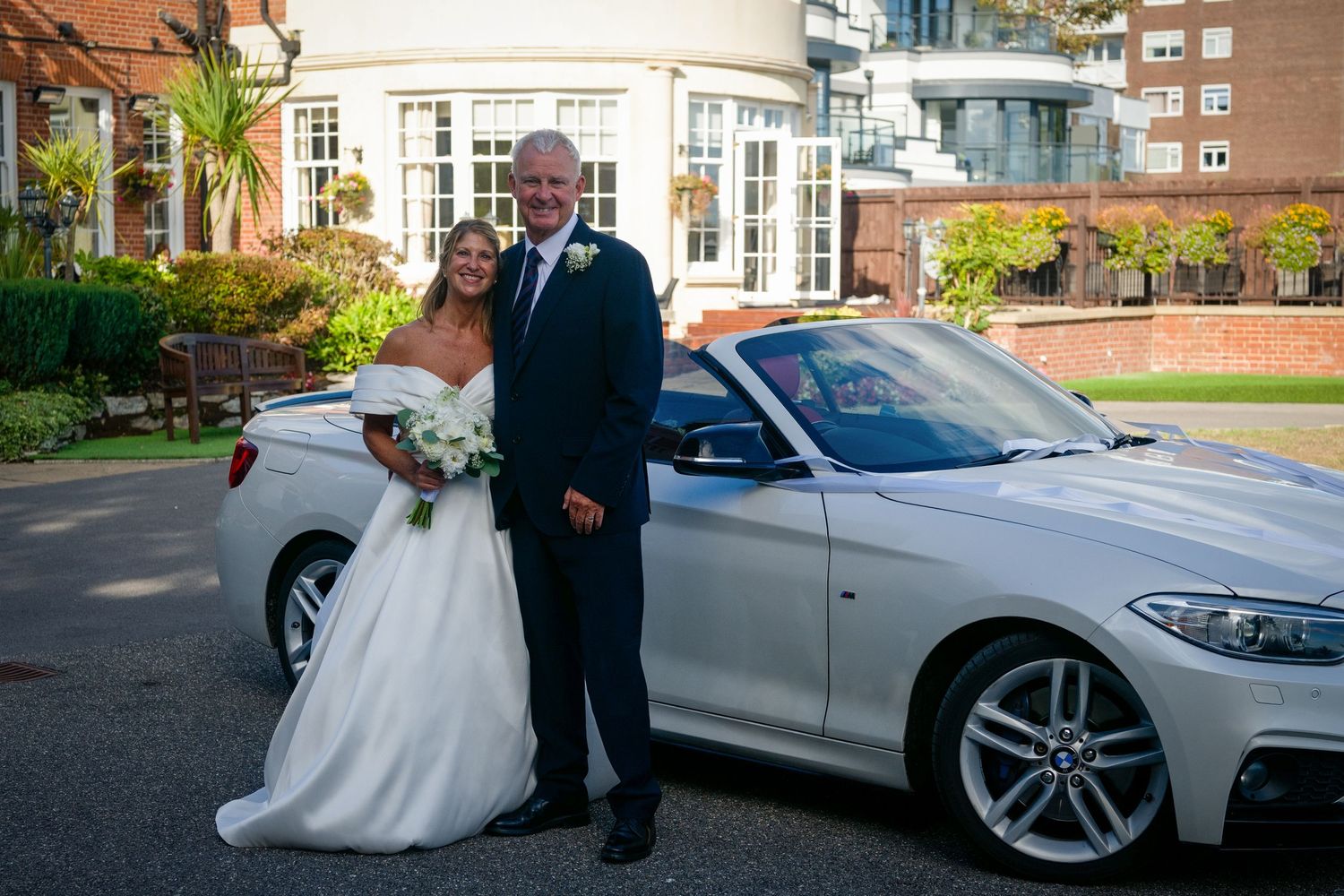 Bride and groom posing beside a white BMW convertible outside an elegant venue on their wedding day.