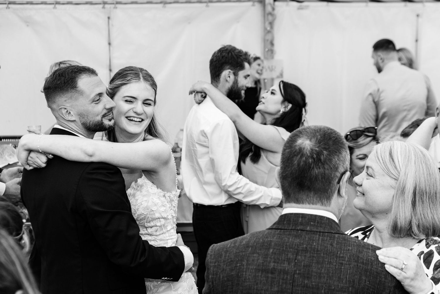 Black and white photo of bride dancing and smiling with guests at wedding reception.