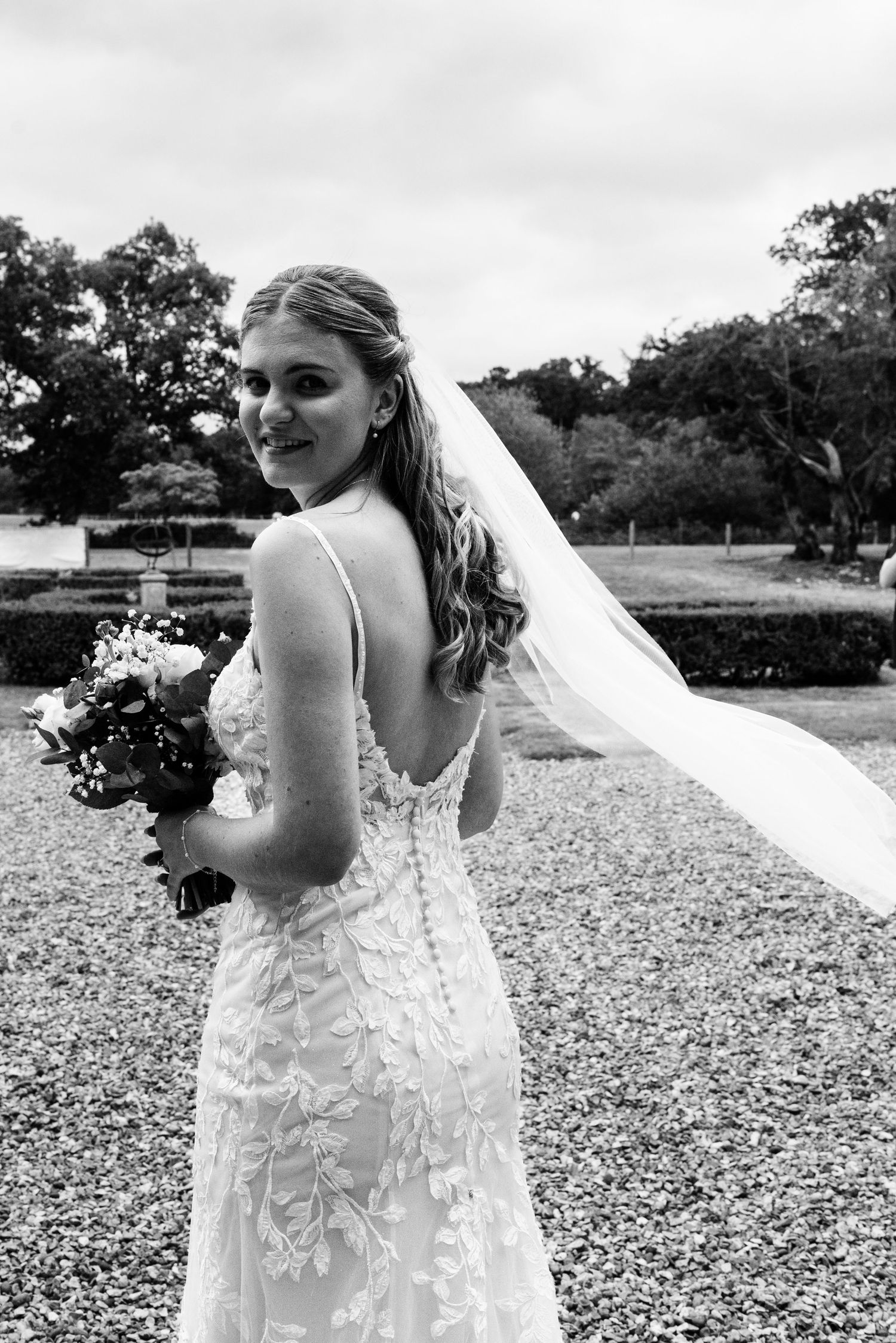 Bride in lace backless wedding dress and veil holds bouquet, smiling over shoulder in black and white photo.
