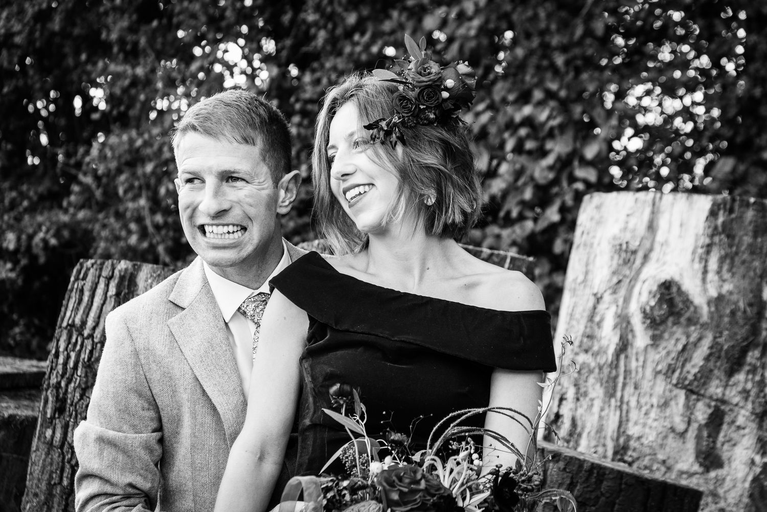 Black and white photo of a smiling couple at a wedding, woman in black dress with floral headpiece holding a bouquet.