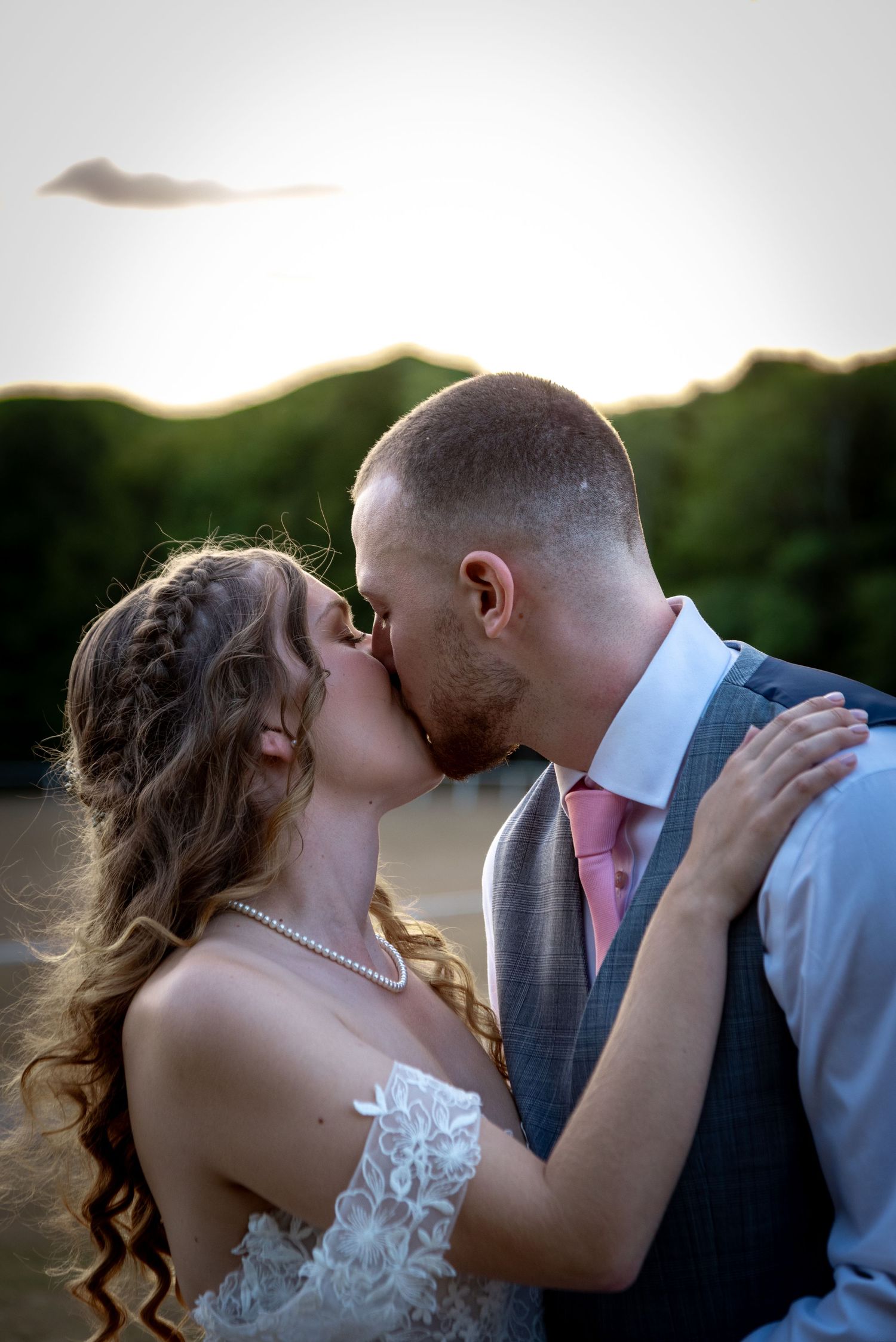 Bride and groom share romantic sunset kiss outdoors, bride in lace dress with pearl necklace.