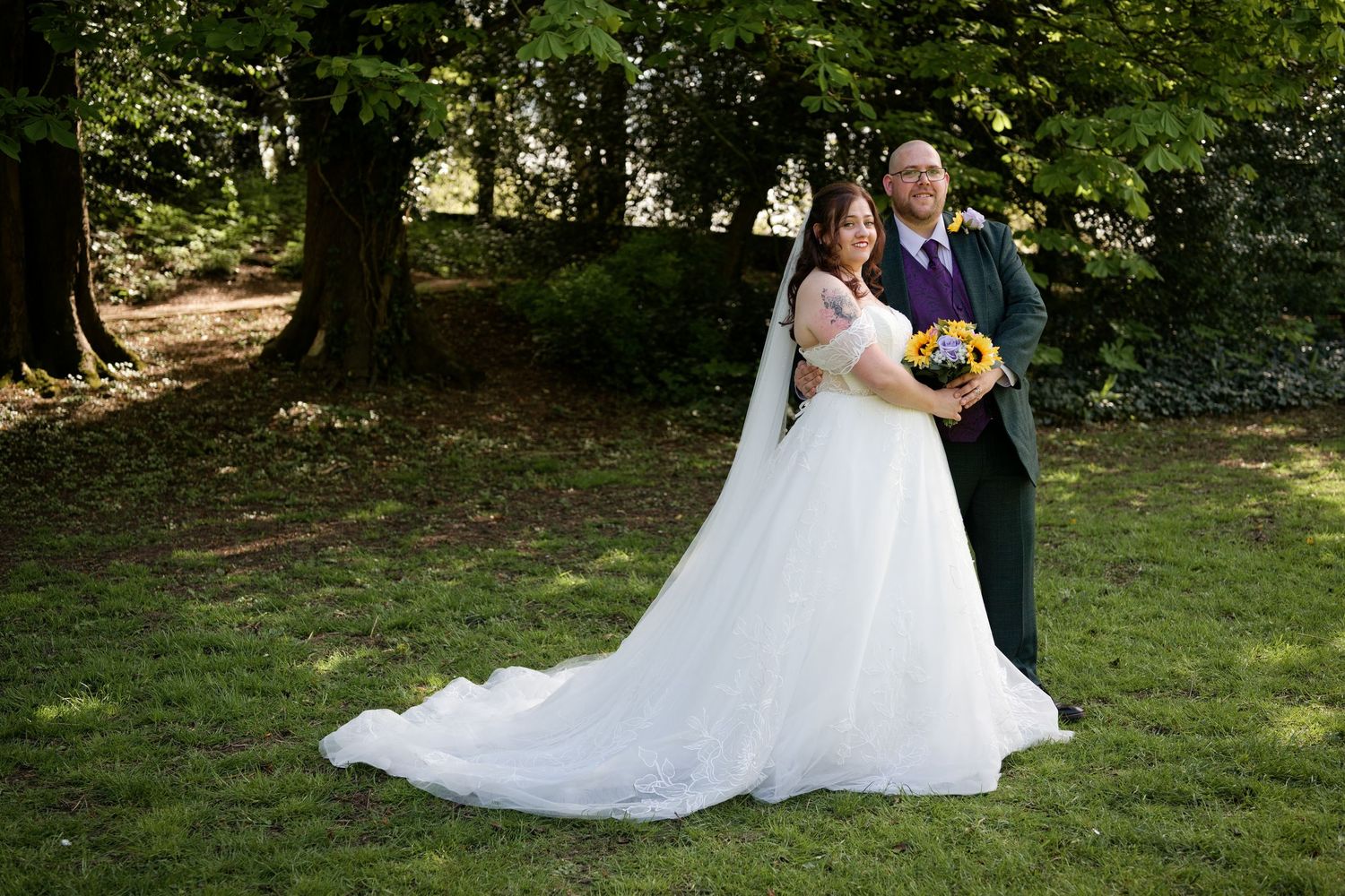 A bride in a white ballgown and groom in a dark suit pose together on a green lawn surrounded by trees.
