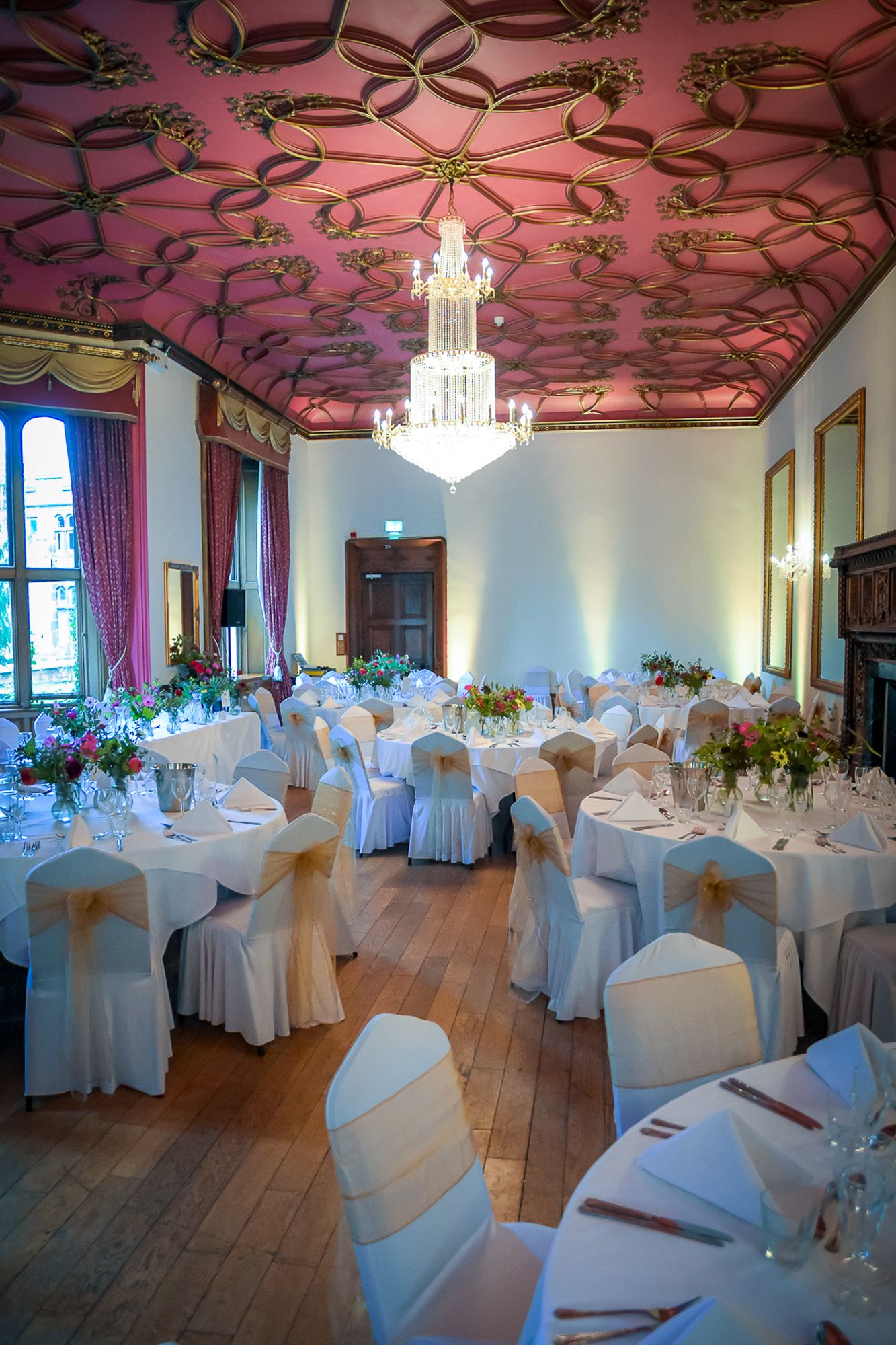 Elegant wedding reception hall with ornate pink ceiling, chandelier, white chair covers, and floral centerpieces on round tables.