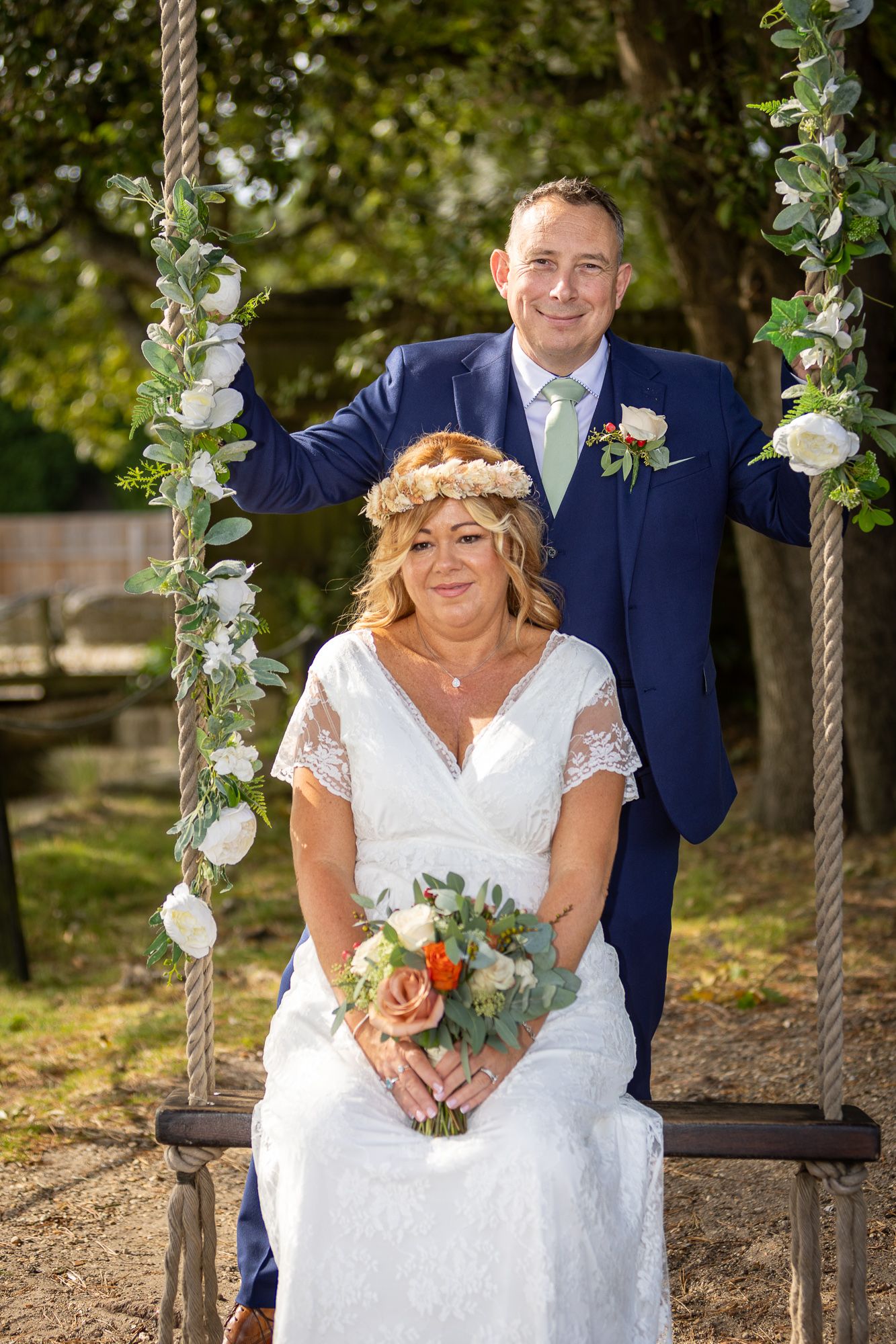 Bride sitting on floral swing holding bouquet, groom standing behind in navy suit, outdoor wedding portrait.