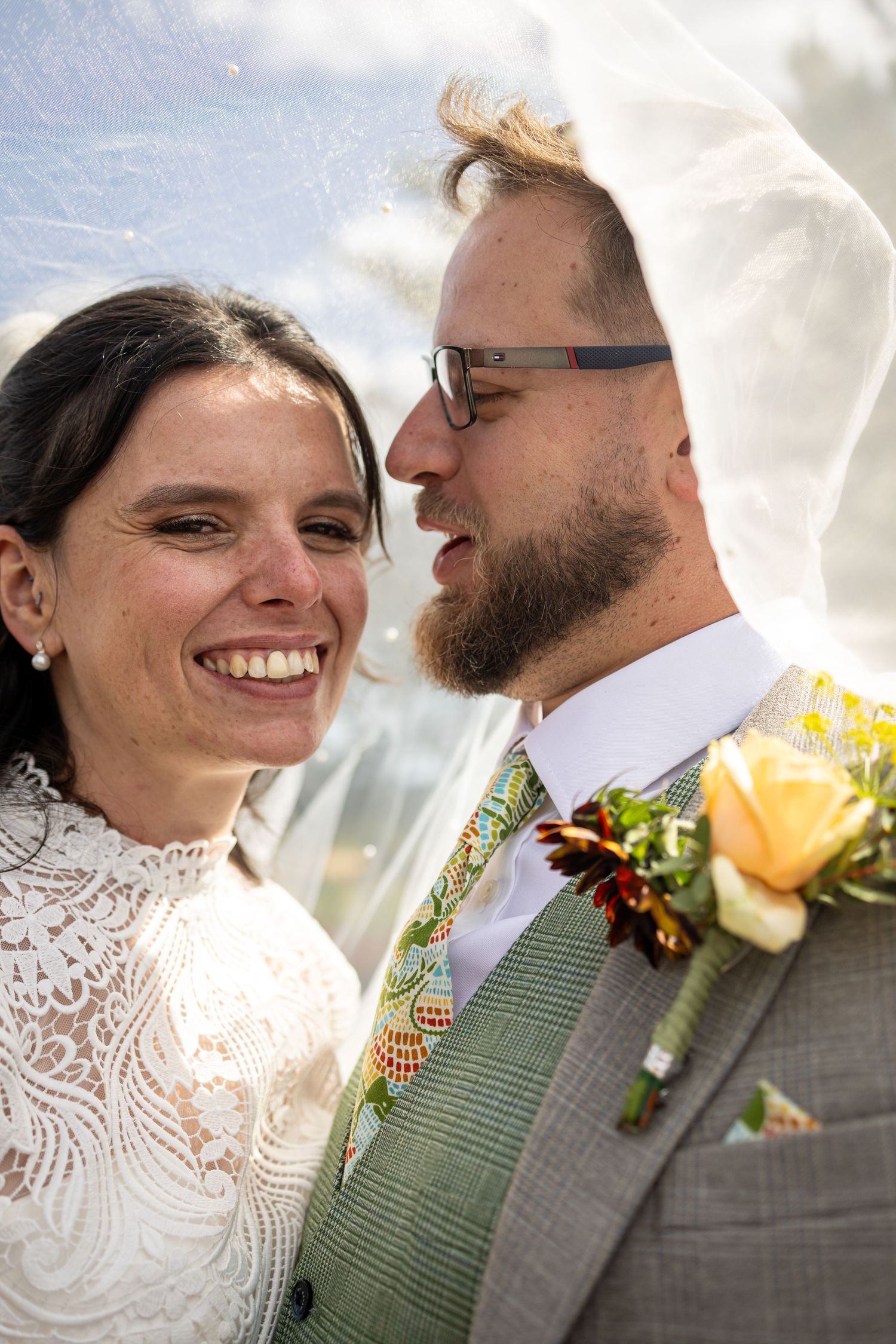 A smiling bride and groom share a romantic moment under a veil, groom wearing glasses and a floral boutonniere.