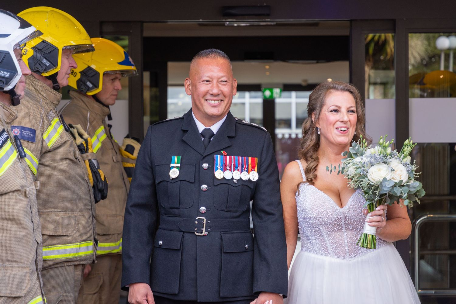 Man in military uniform with medals walks beside a bride holding a bouquet, with firefighters behind.