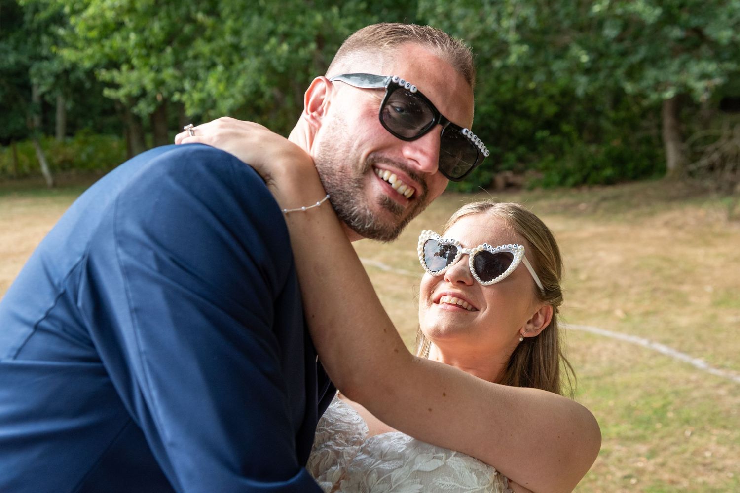 A smiling groom in a navy suit and bride in heart-shaped sunglasses share a joyful embrace outdoors.