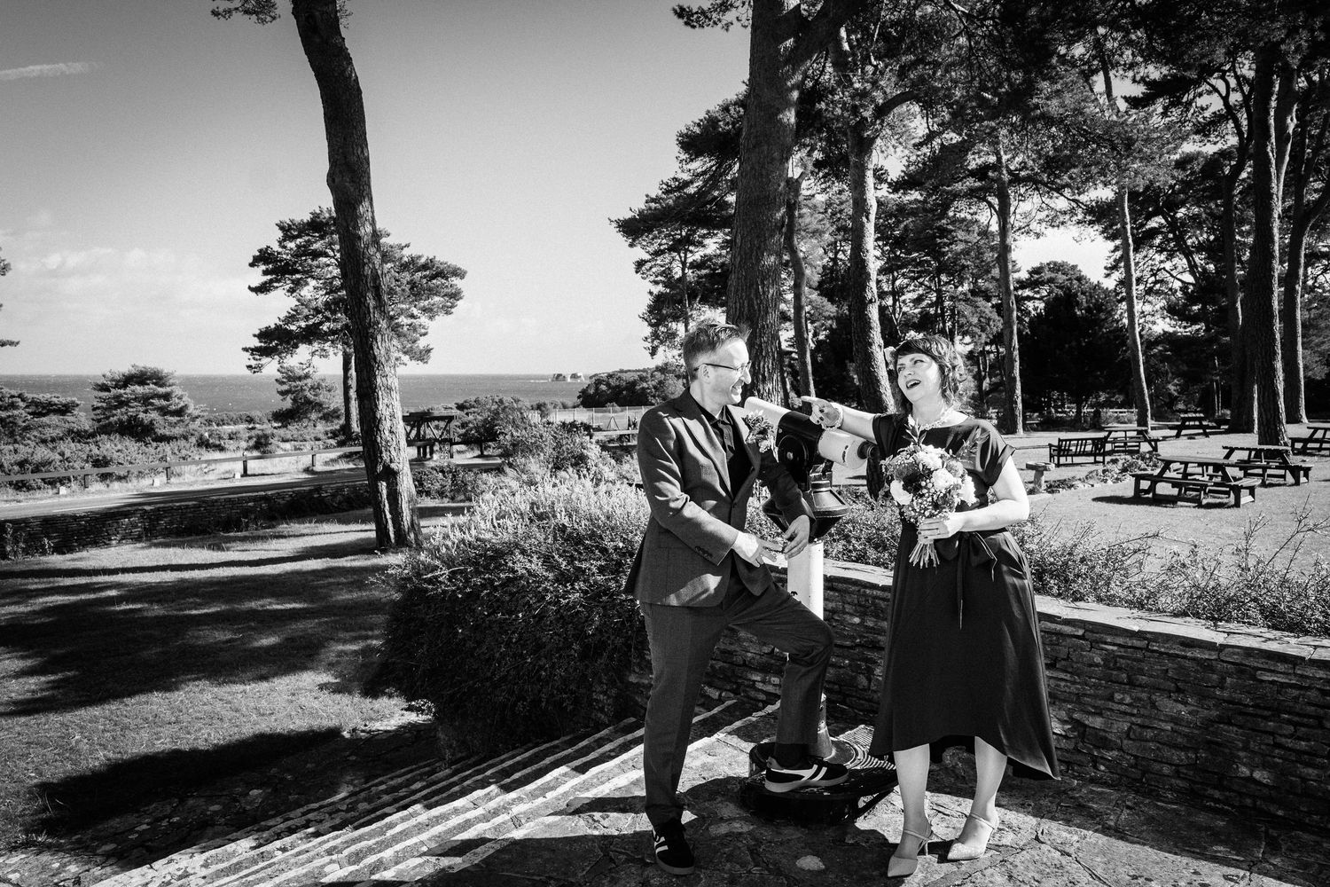 Black and white photo of a couple dancing outdoors at a woodland venue with picnic tables and trees in background.