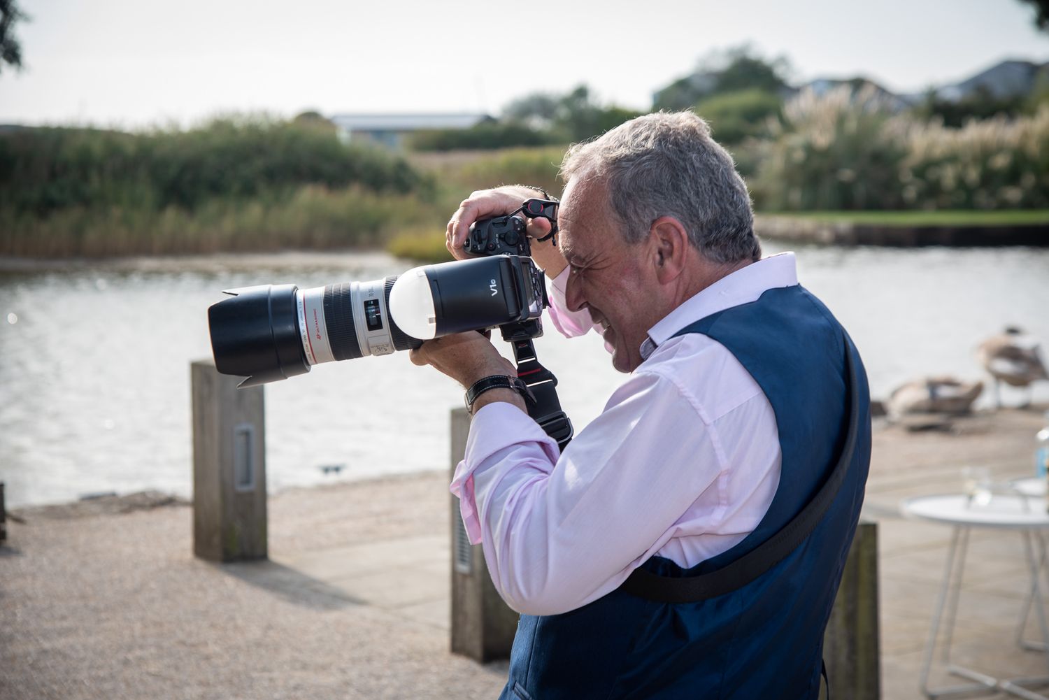 A person wearing a blue vest and white shirt photographs wildlife with a telephoto lens on a sunny day at the beach.