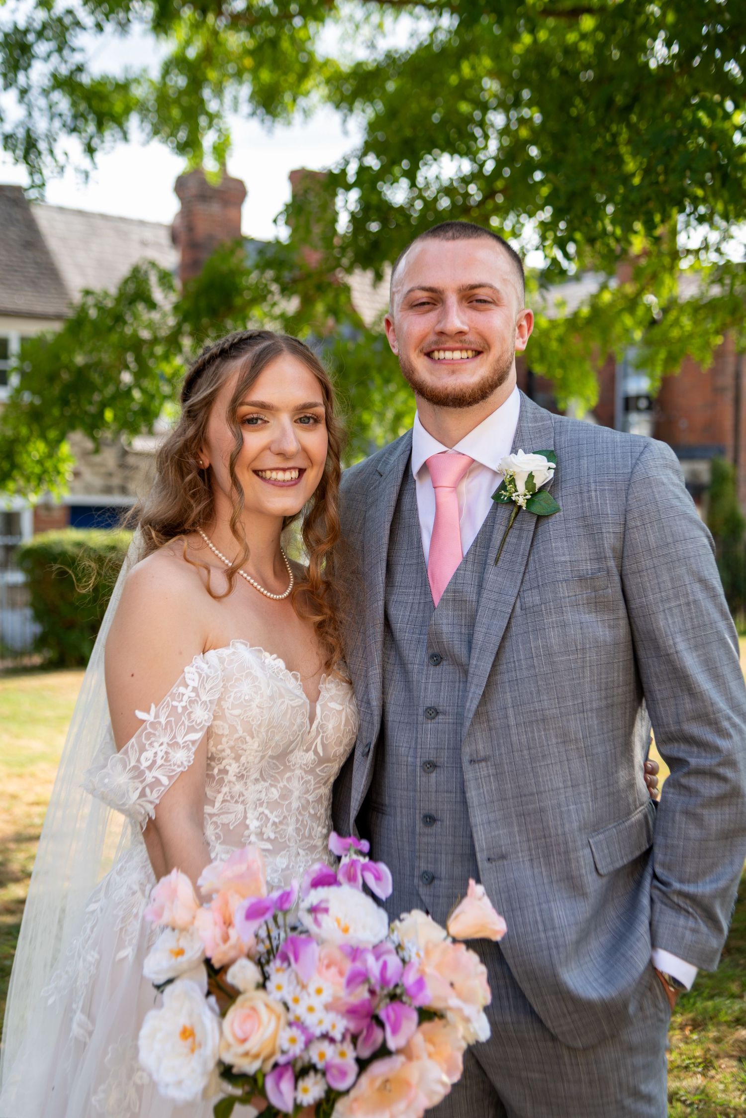 Smiling bride holding a colorful bouquet stands with her groom in a grey suit and pink tie under a leafy tree.