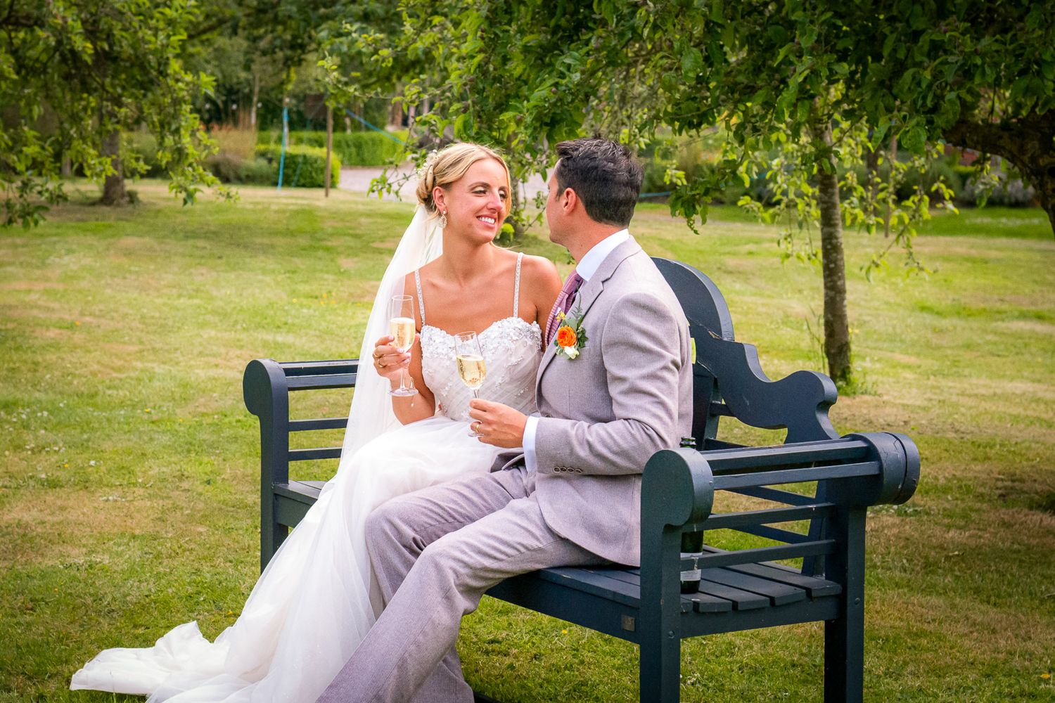 Bride and groom sitting on a dark green bench in a sunny garden, sharing champagne and smiling at each other.