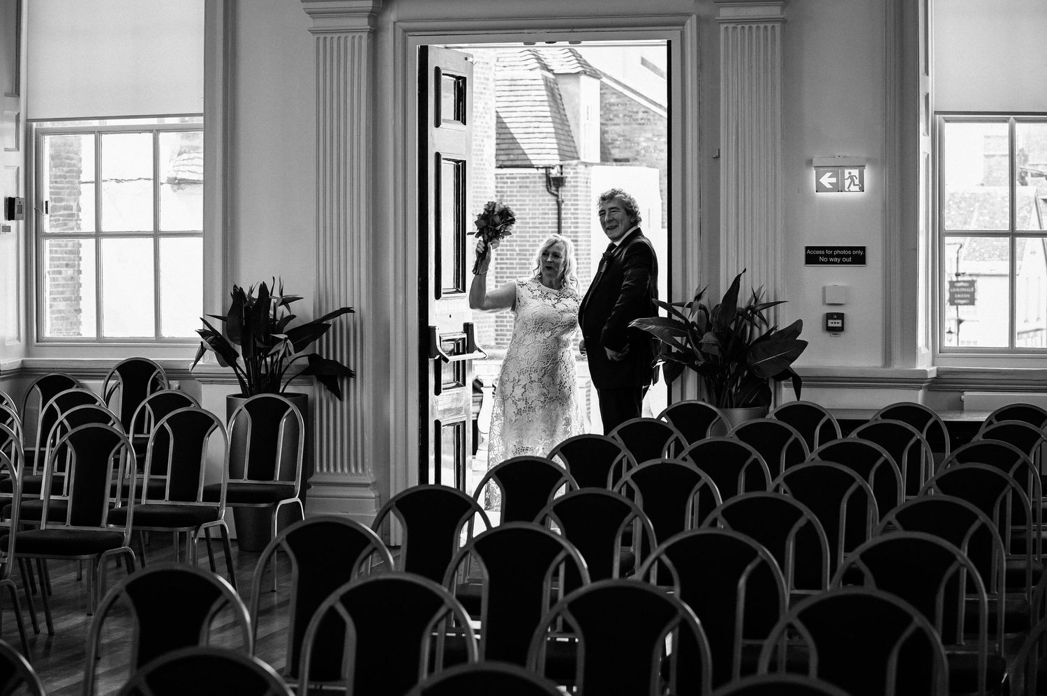 Black and white photo of a bride and groom standing at the entrance of an elegant wedding ceremony venue with rows of chairs.