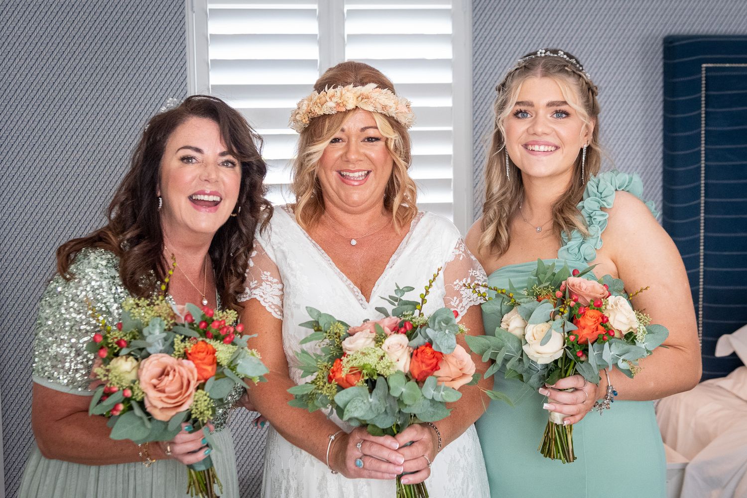 Bride in white dress with two bridesmaids in mint and silver, holding colorful bouquets, smiling indoors.